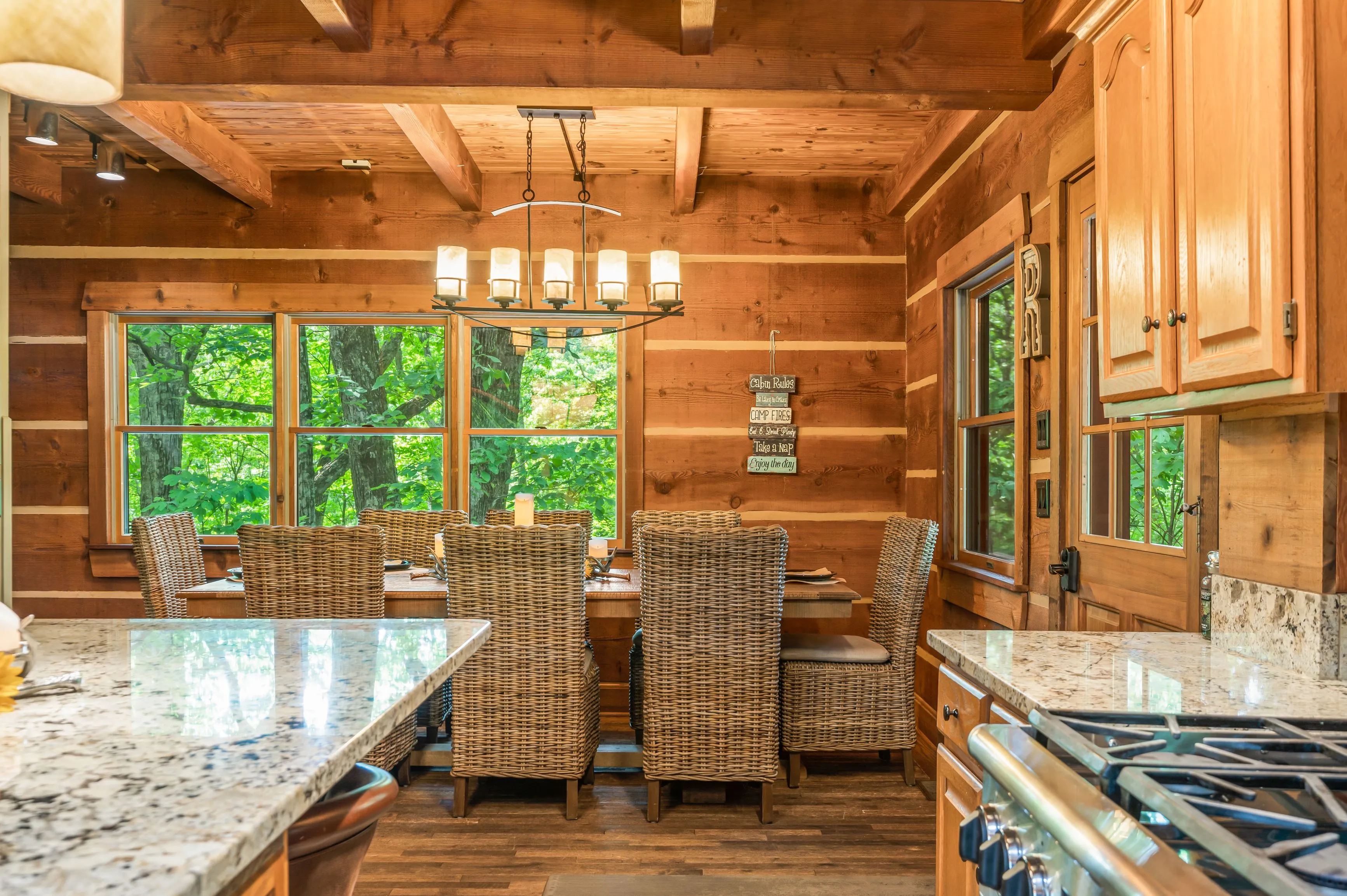 Rustic wooden kitchen interior with a dining table set for six, wicker chairs, granite countertops, and a forest view through large windows.