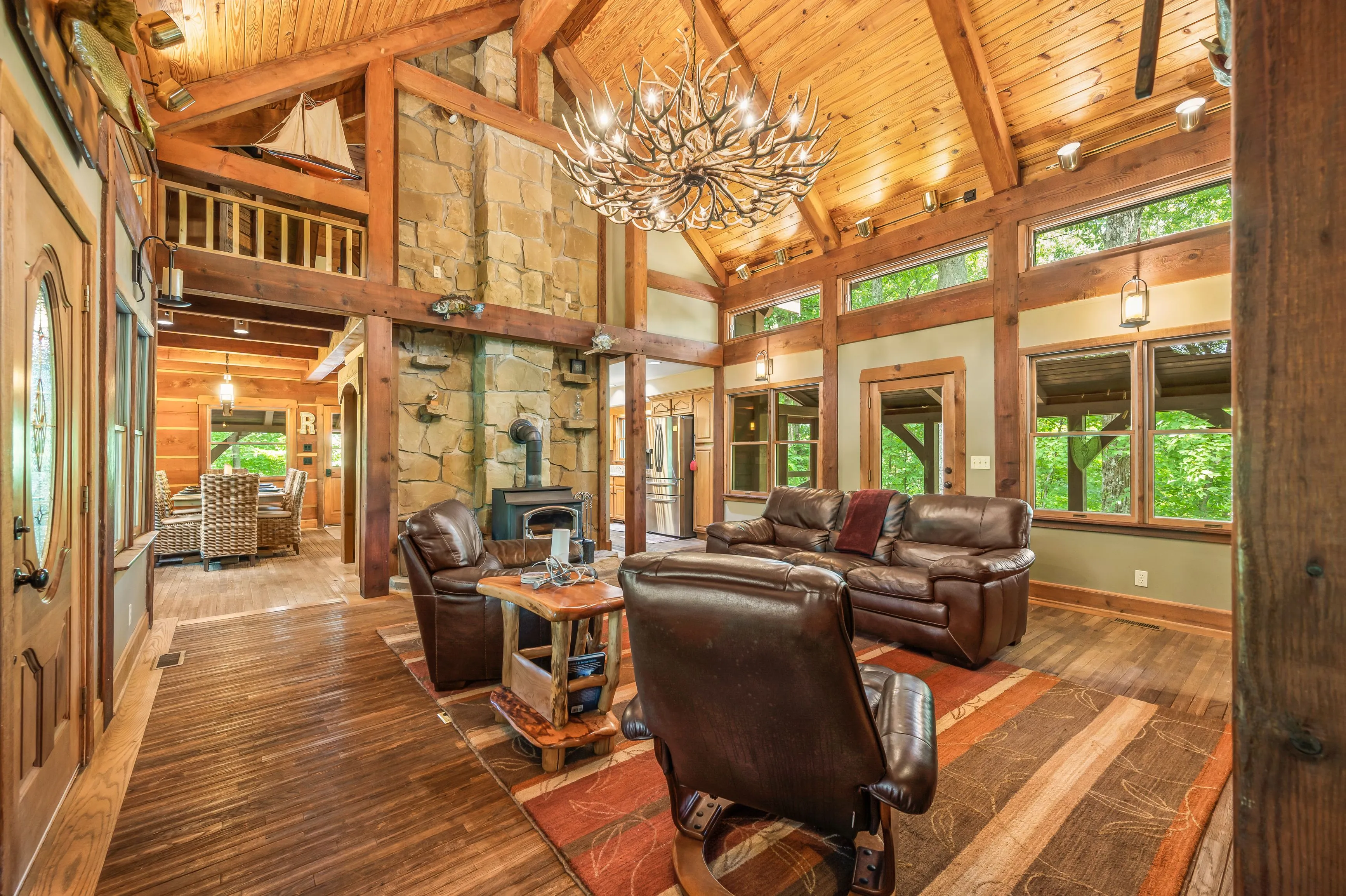 Spacious and cozy rustic living room interior with high vaulted ceiling, leather furniture, stone fireplace with a wood-burning stove, antler chandelier, and large windows overlooking greenery.