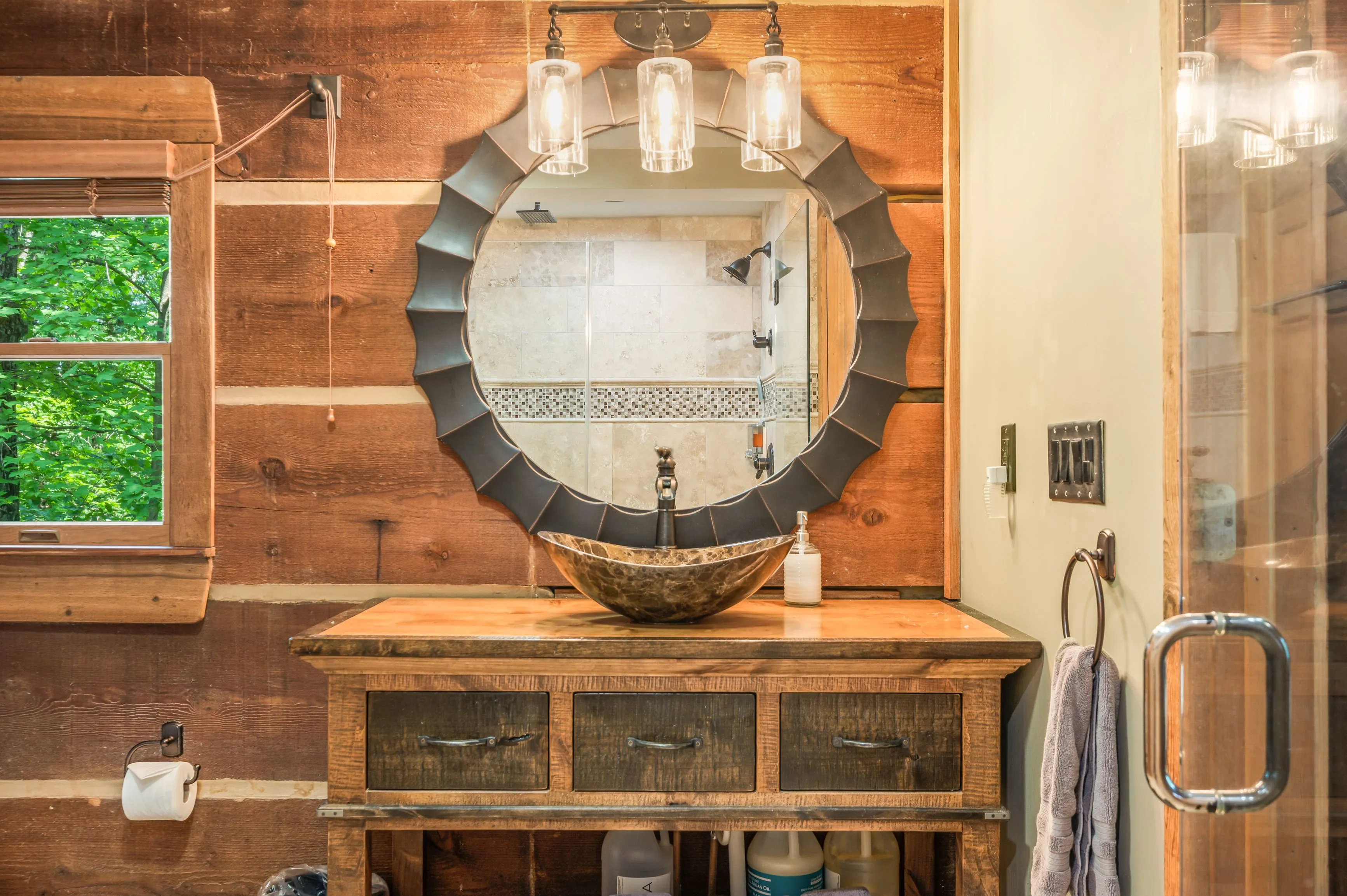 Rustic bathroom interior with wooden vanity, stone vessel sink, round mirror with industrial-style sconces, shower visible in mirror reflection, and natural light from a window.