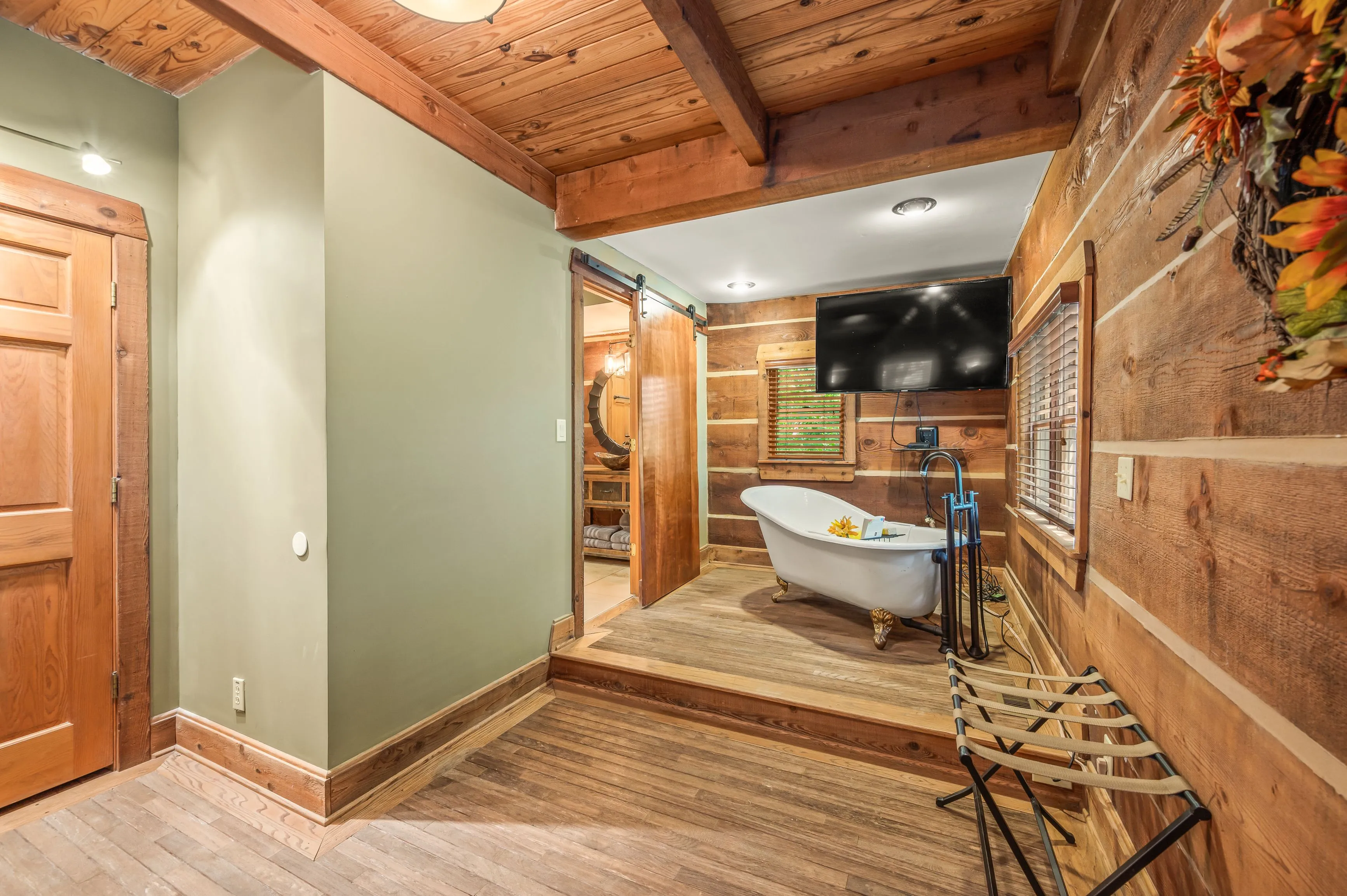 Rustic-style bathroom interior with a freestanding clawfoot bathtub, wooden walls and floors, and sliding barn door leading to a separate room.