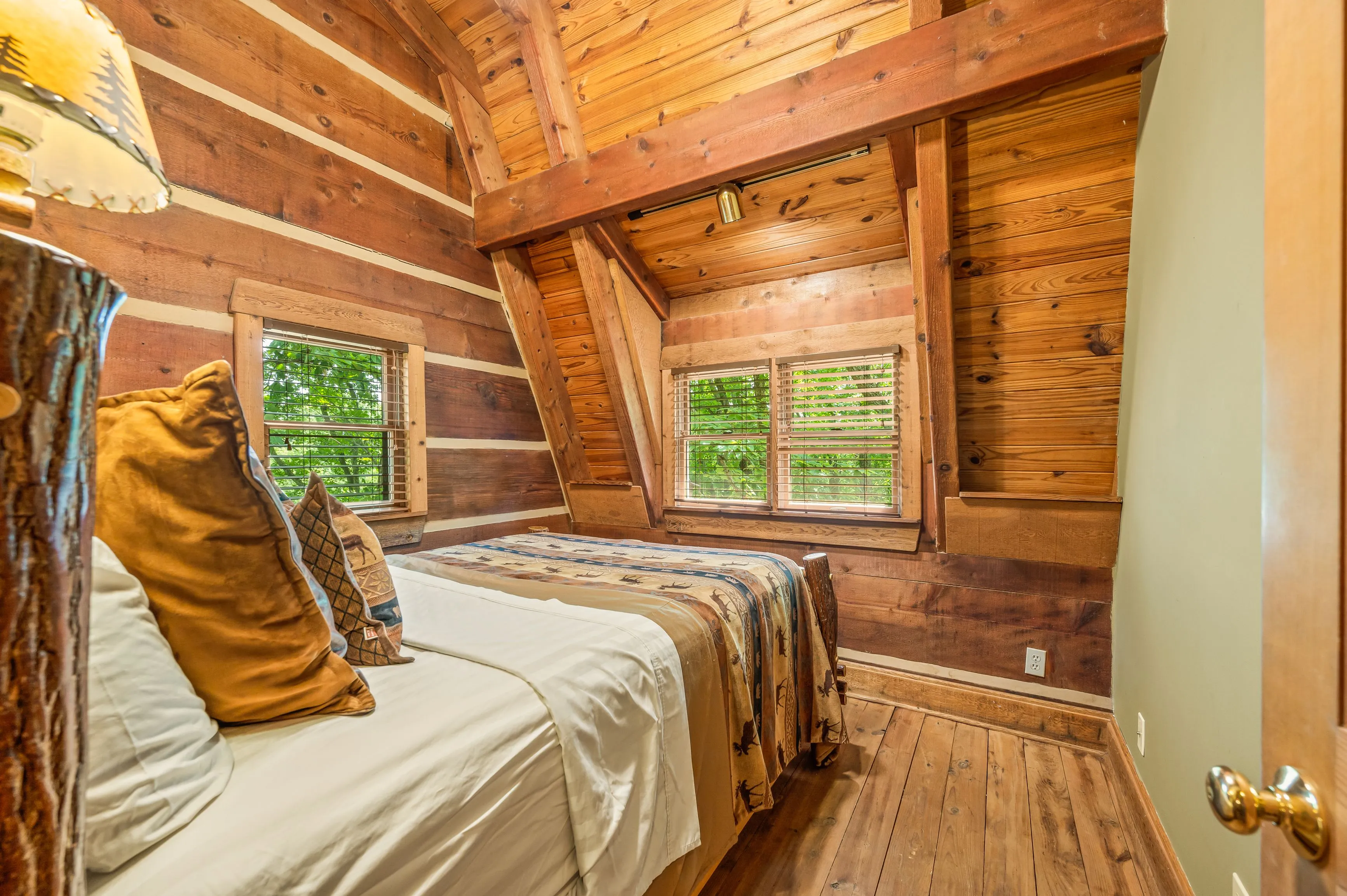 Cozy wooden cabin bedroom interior with a bed covered in a patterned quilt, rustic wood paneled walls, and windows overlooking greenery.