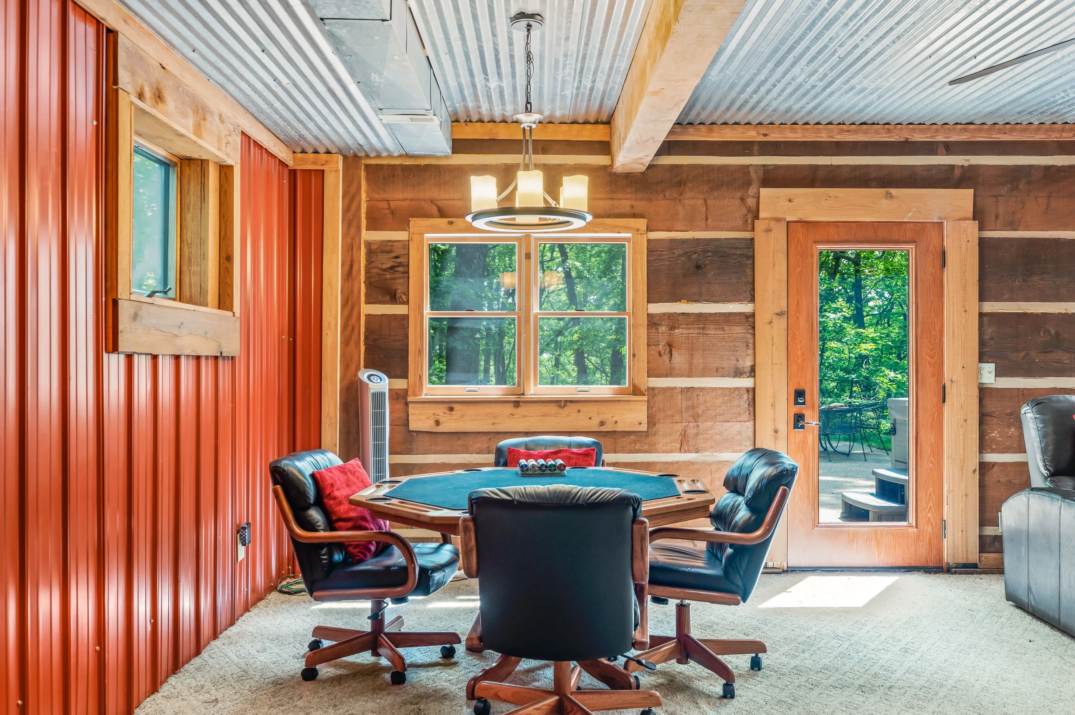 Rustic home office with corrugated metal ceiling, red accent wall, log cabin walls, a hexagonal wooden desk with leather chairs, and windows looking out to greenery.