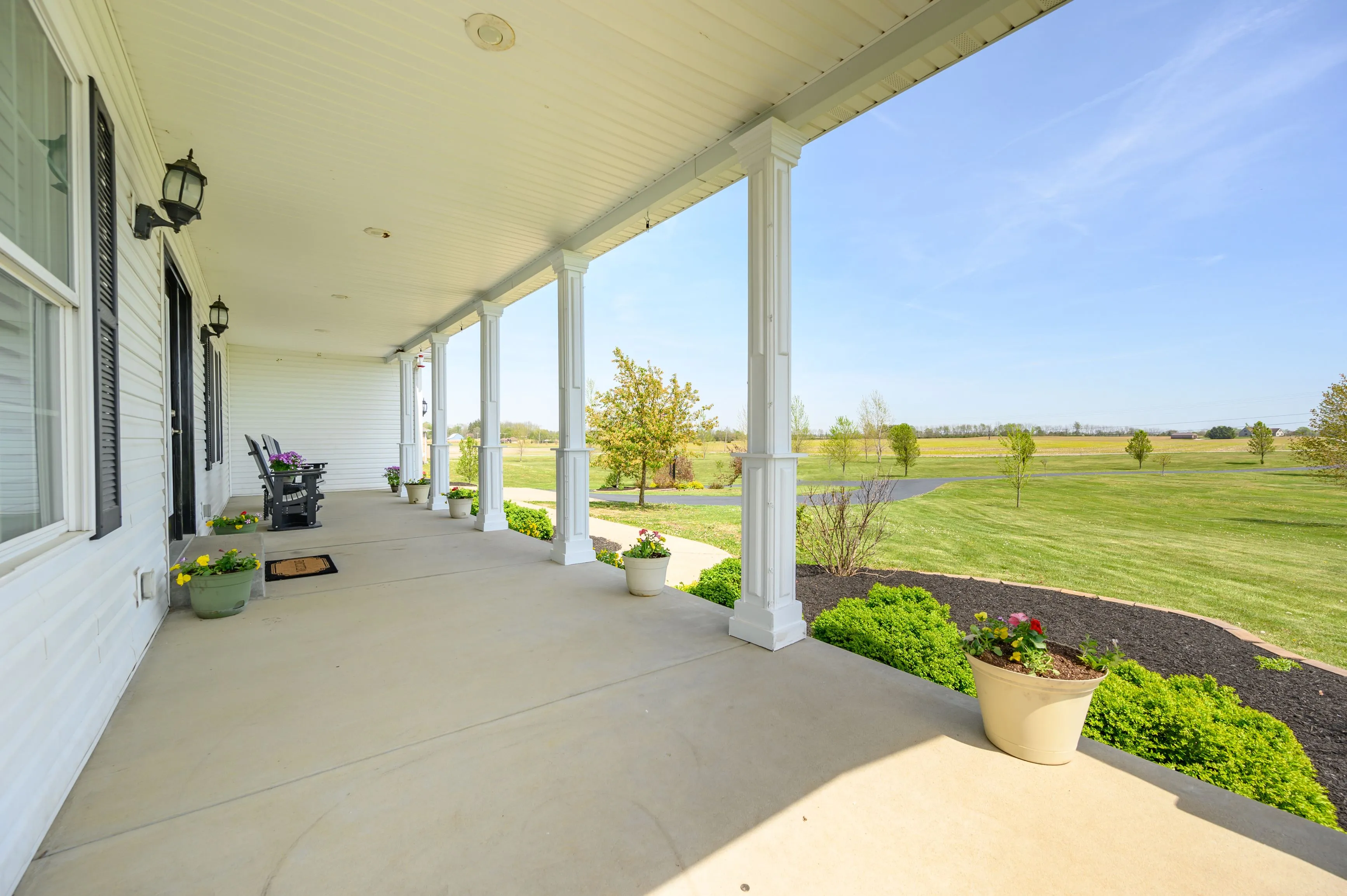 Spacious porch of a country house with columns, overlooking a vast lawn and clear blue sky.