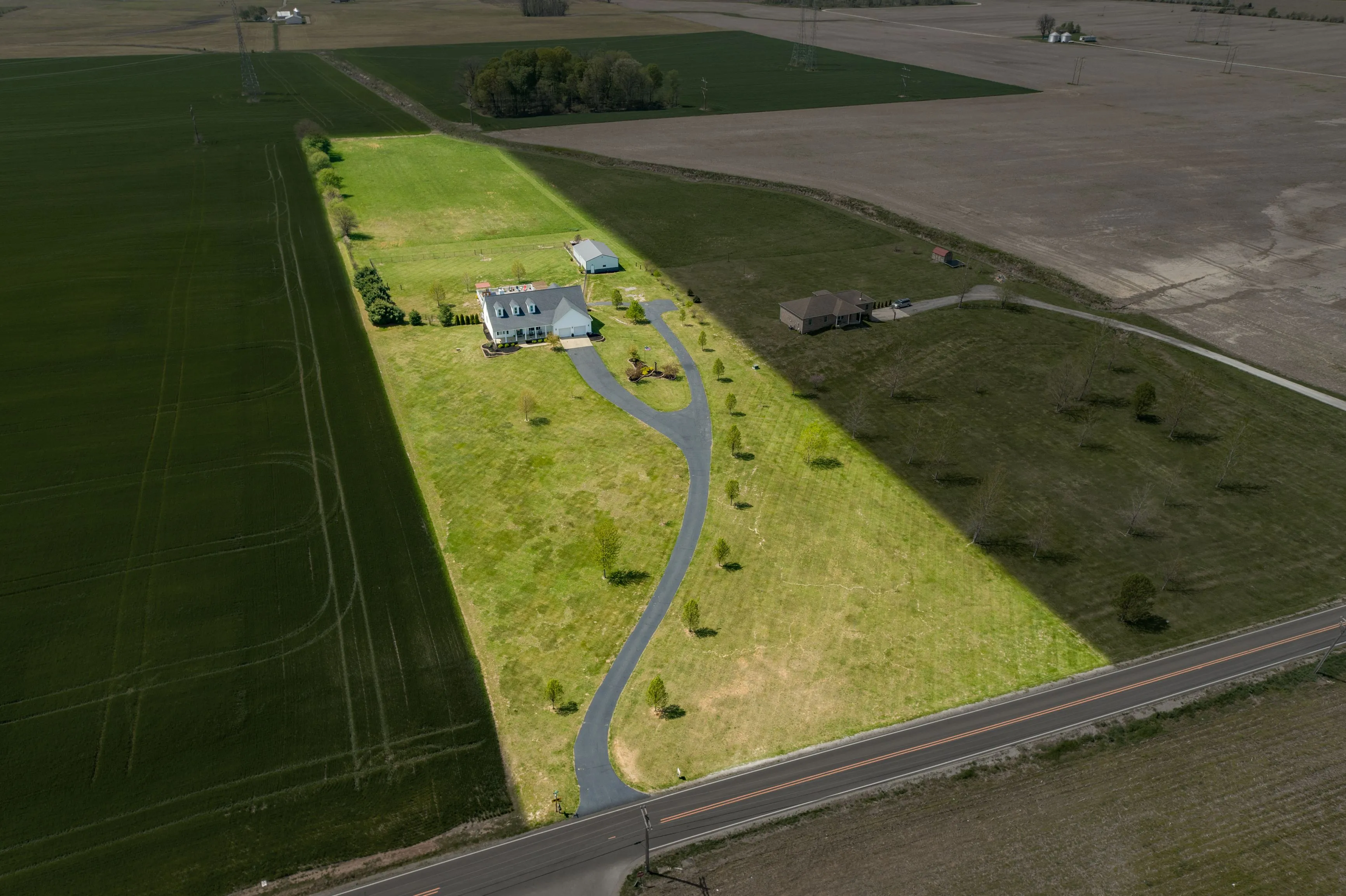 Aerial view of a green rectangular land plot with a curvy path leading to a house, surrounded by fields with varying shades of brown.