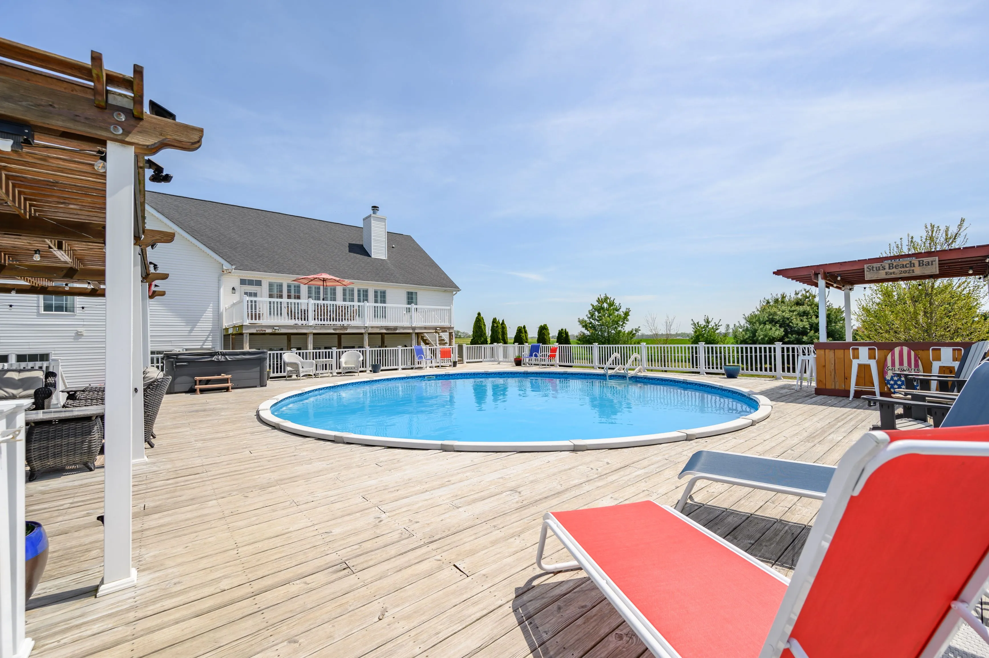 Spacious backyard with a large oval swimming pool, wooden deck, red sun loungers, and a two-story house in the background under a clear blue sky.