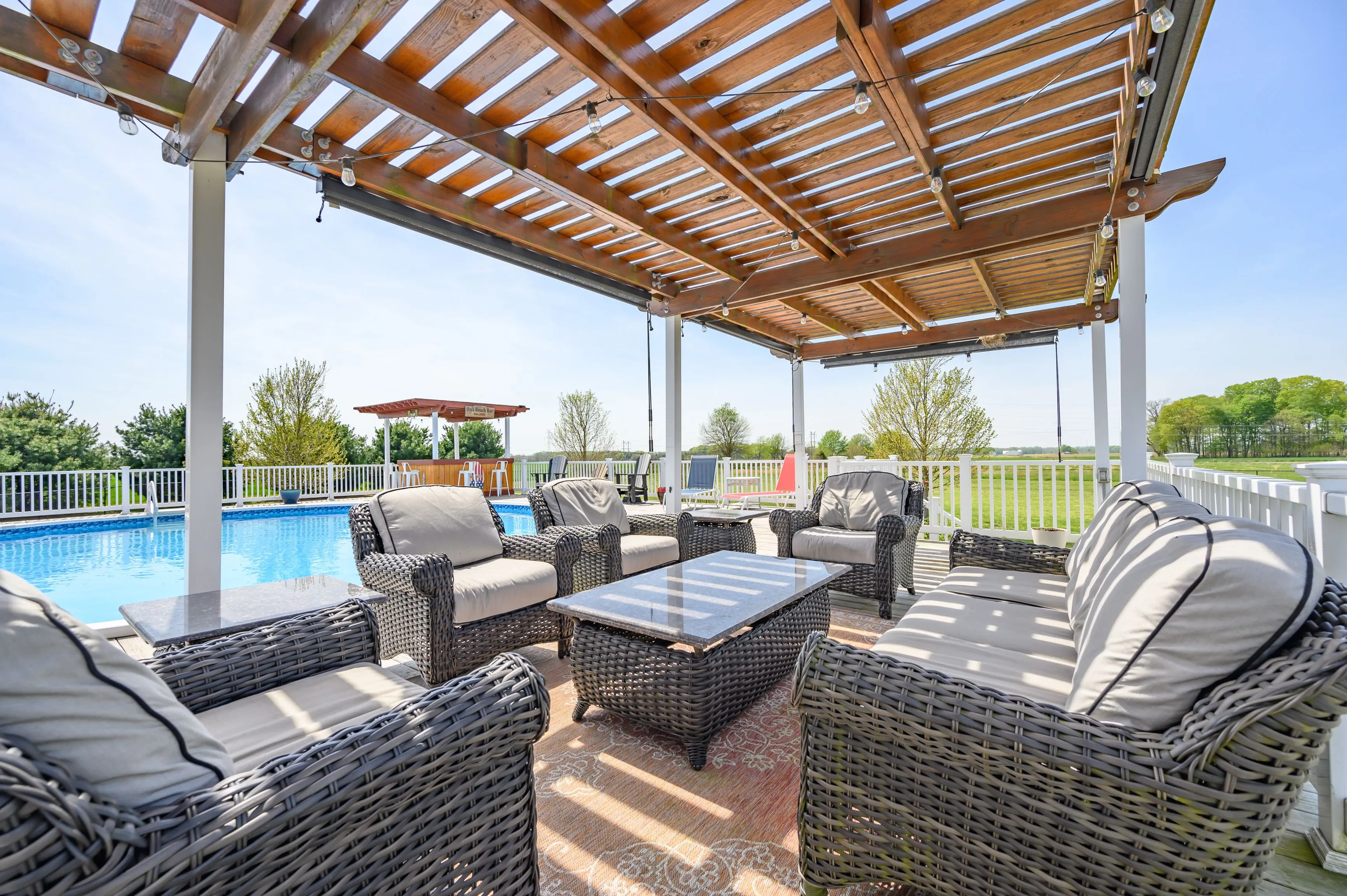 Outdoor patio area with rattan furniture under a pergola, overlooking a swimming pool on a sunny day.