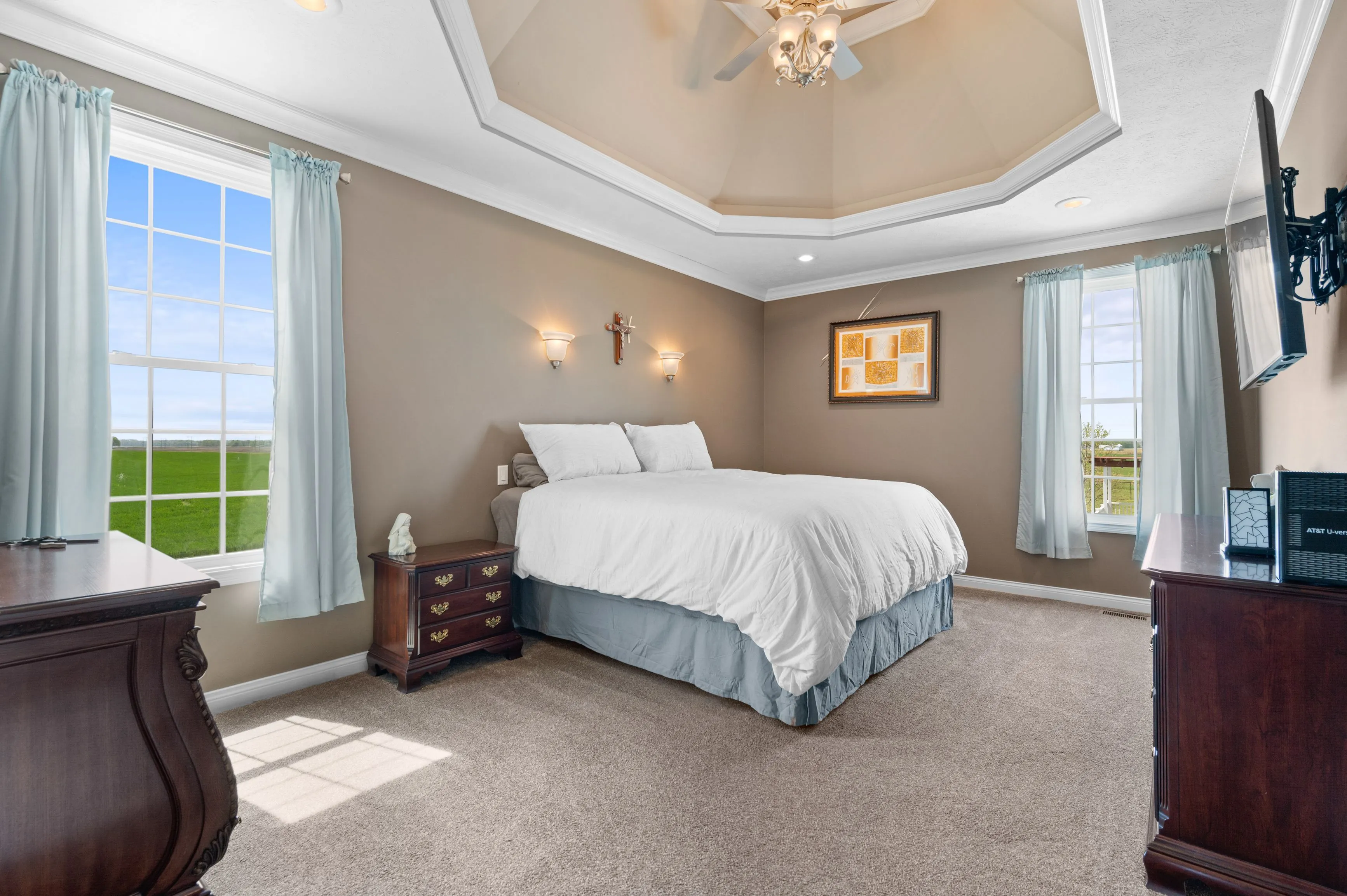 Elegant bedroom with a queen-sized bed, wooden furniture, and a tray ceiling with a ceiling fan.