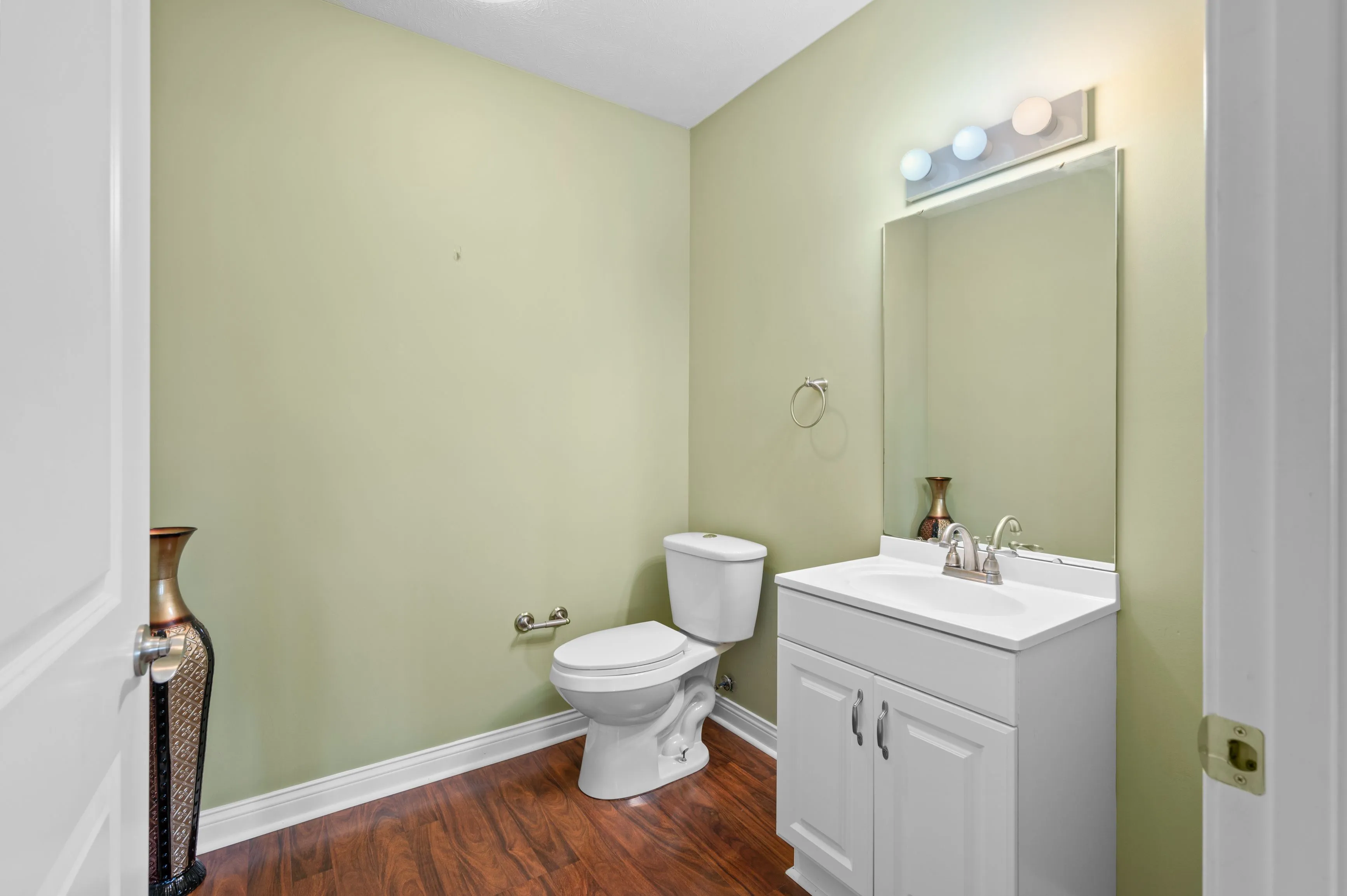 Small bathroom with light green walls, white fixtures, wooden floor, and vanity mirror with lights.