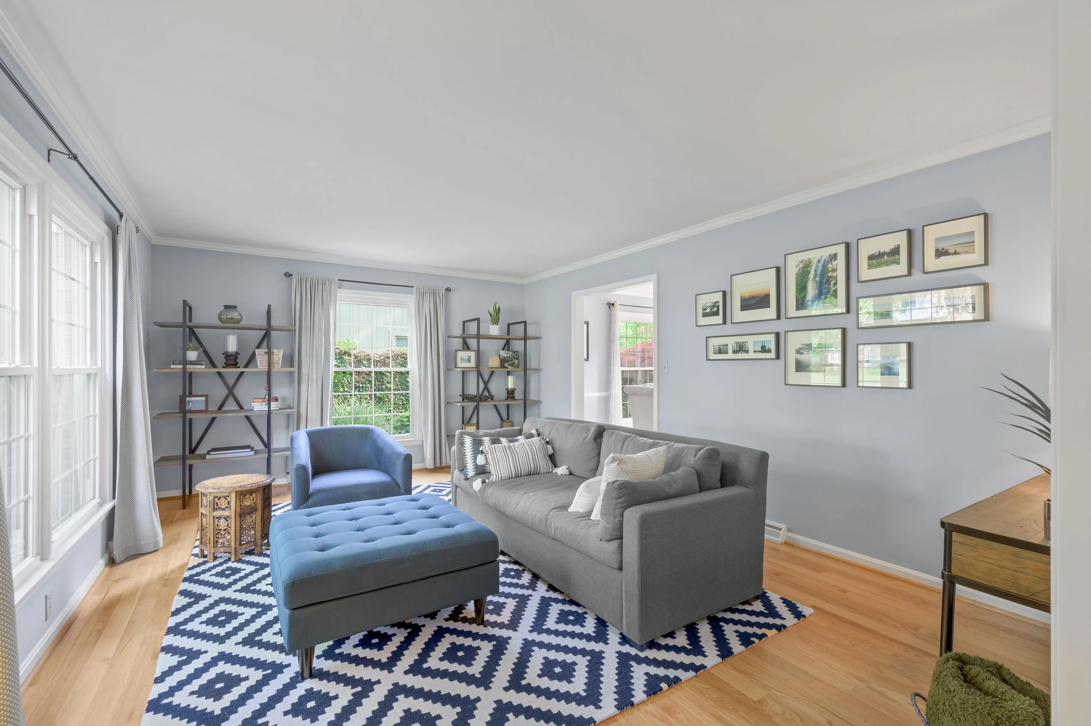 Bright and spacious living room with hardwood floors, a blue patterned area rug, grey sofa, blue armchair, and a wall decorated with framed pictures.
