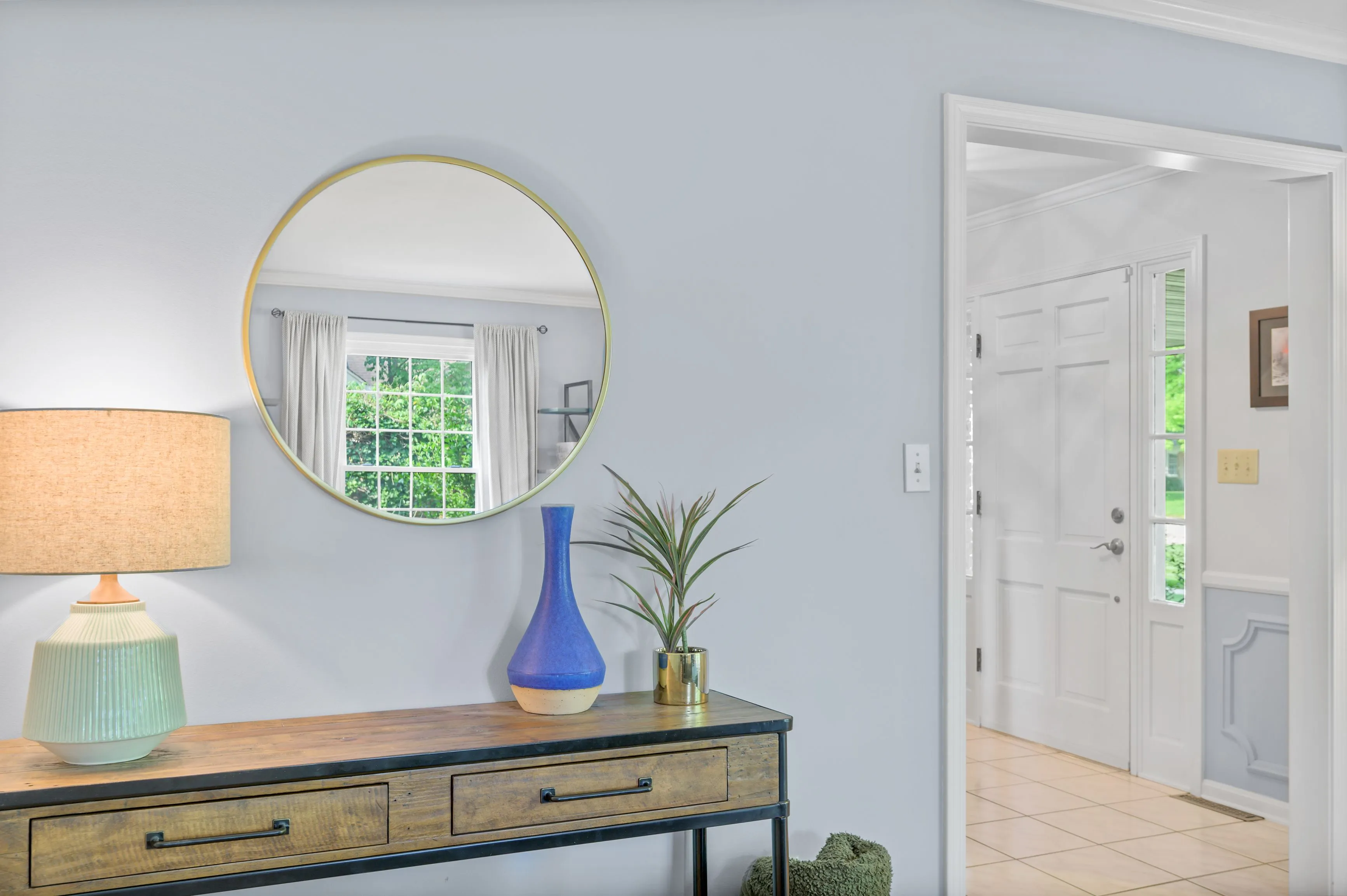 A bright entryway featuring a round mirror above a console table with a lamp and vase, with a view into another room through an open door.