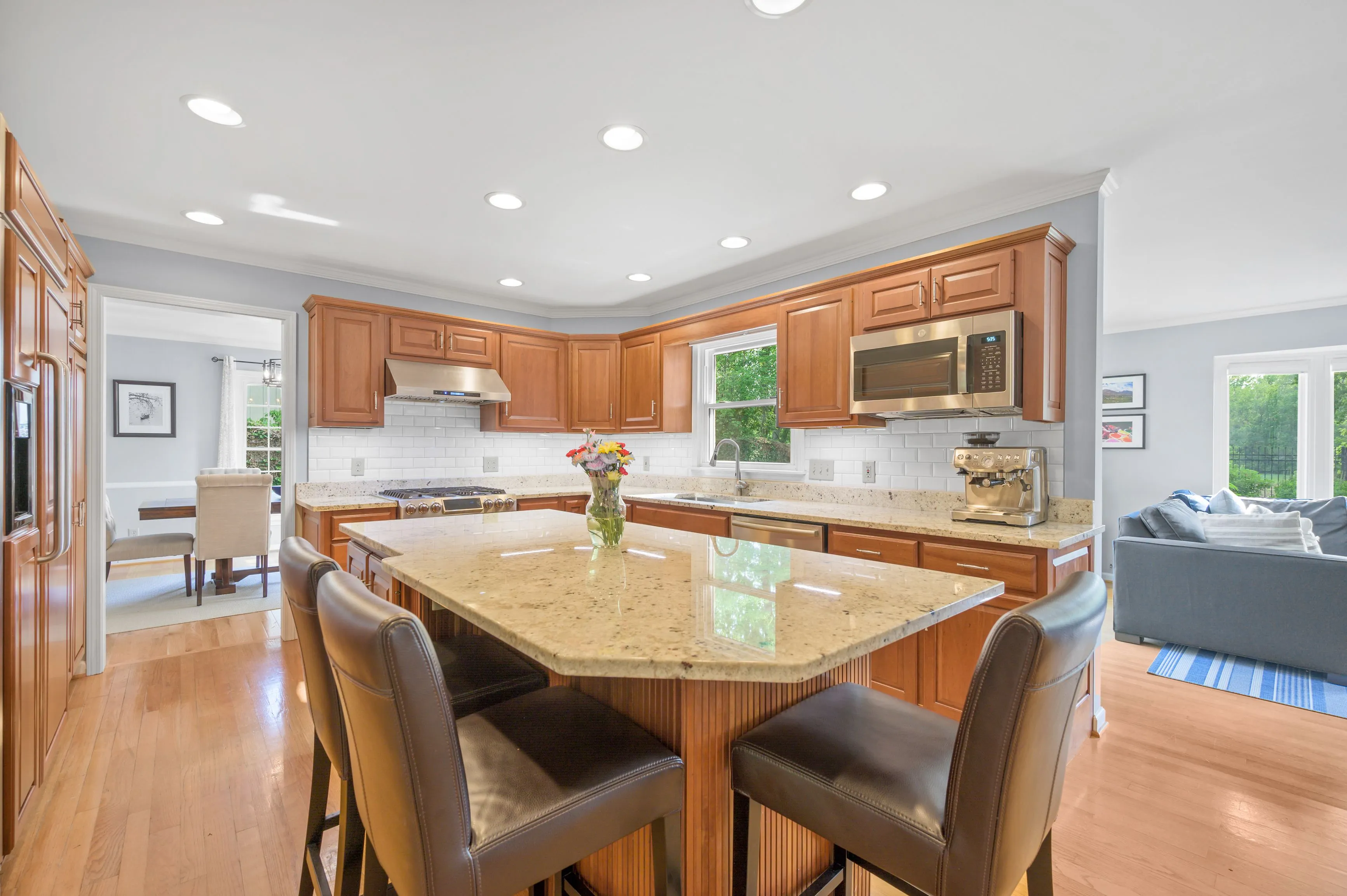 Spacious kitchen interior with wood cabinetry, granite countertops, and modern appliances, connected to a bright dining area with a glass door leading to the outside.