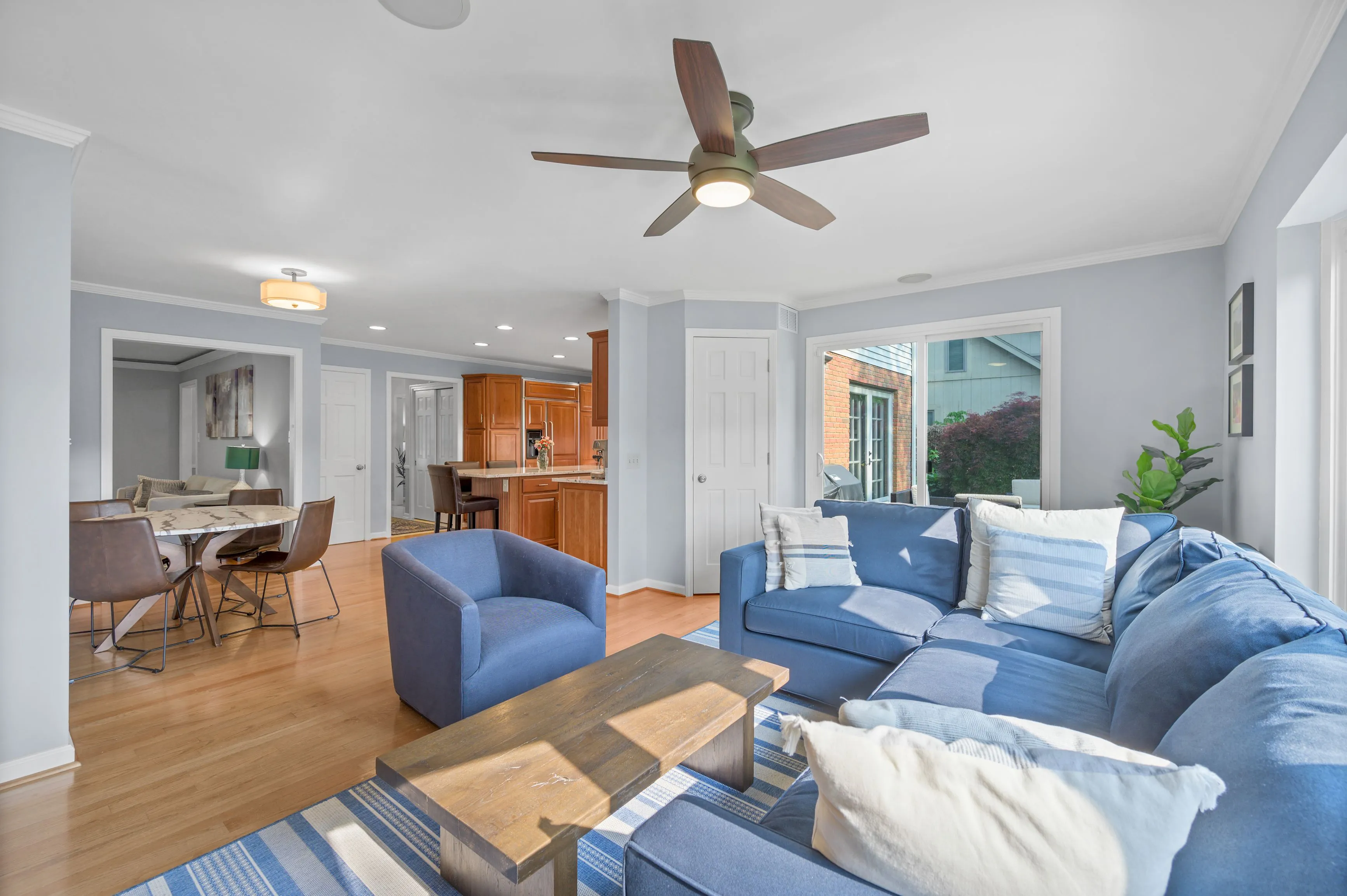 Bright cozy living room with blue sofas, white walls, hardwood floors, and a ceiling fan, adjacent to a dining area and kitchen.