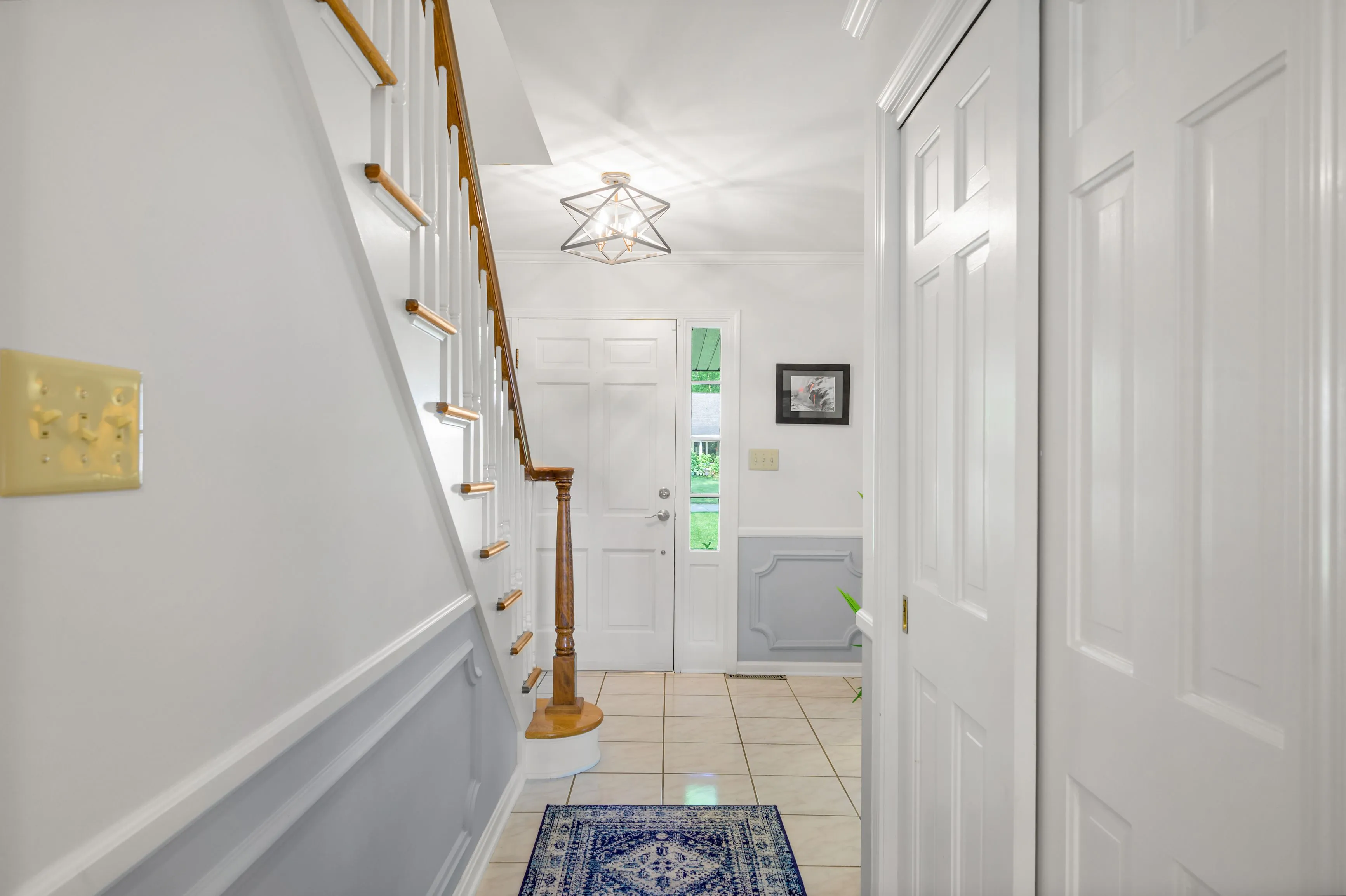 Narrow hallway in a home with white walls, a staircase on the left, and a blue decorative rug on the floor leading to other rooms.