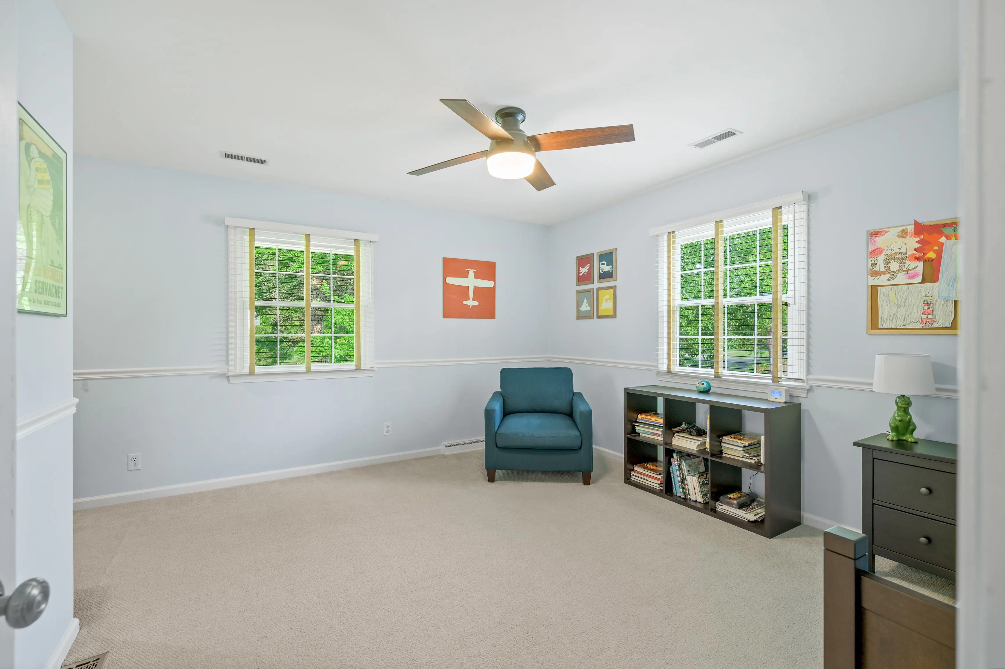 Bright empty room with beige carpet, one blue armchair, ceiling fan, and office shelves by the window with greenery outside.