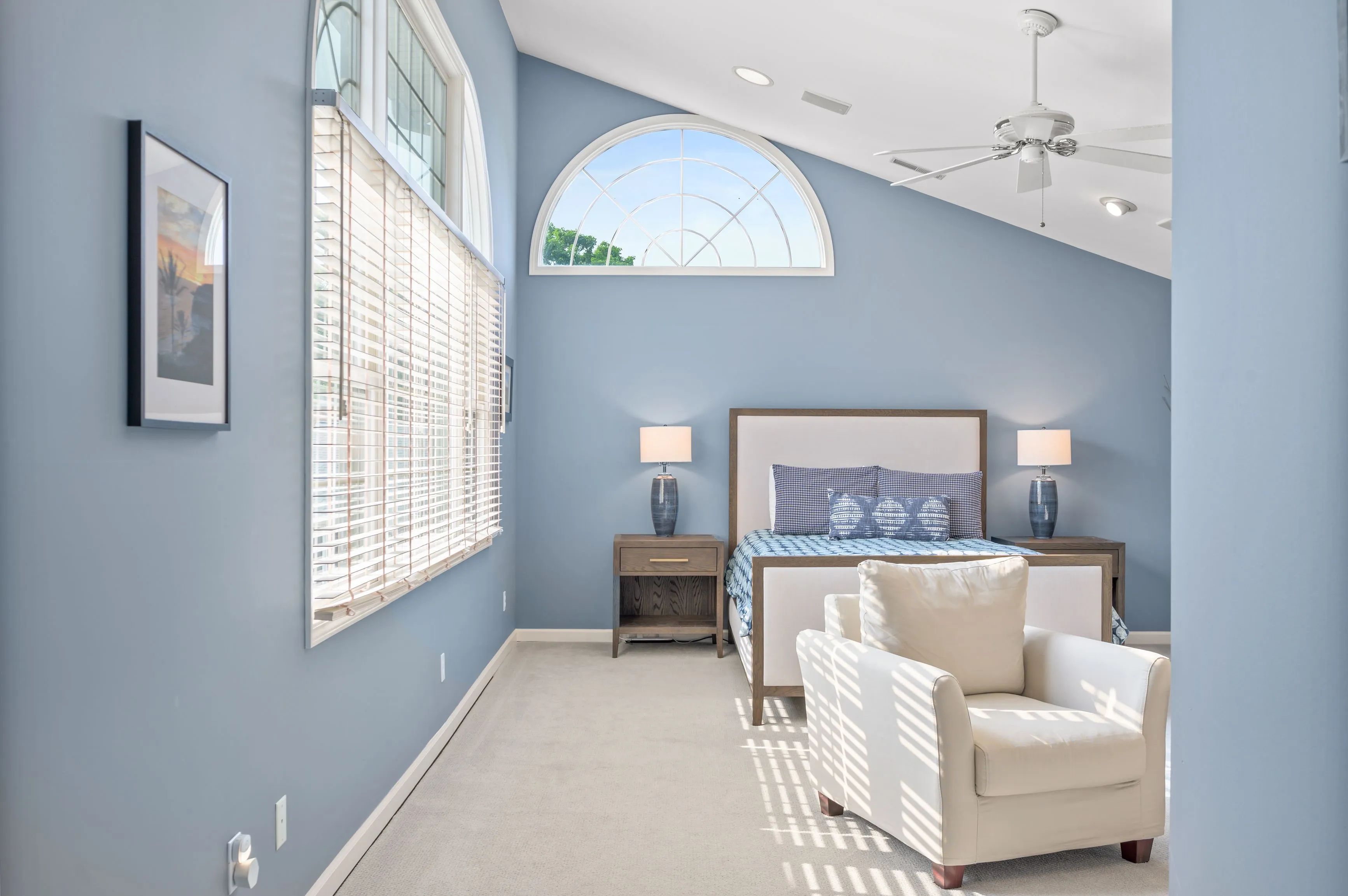 Elegant bedroom interior with light blue walls, large bed with headboard, armchair, and high ceiling with fan.