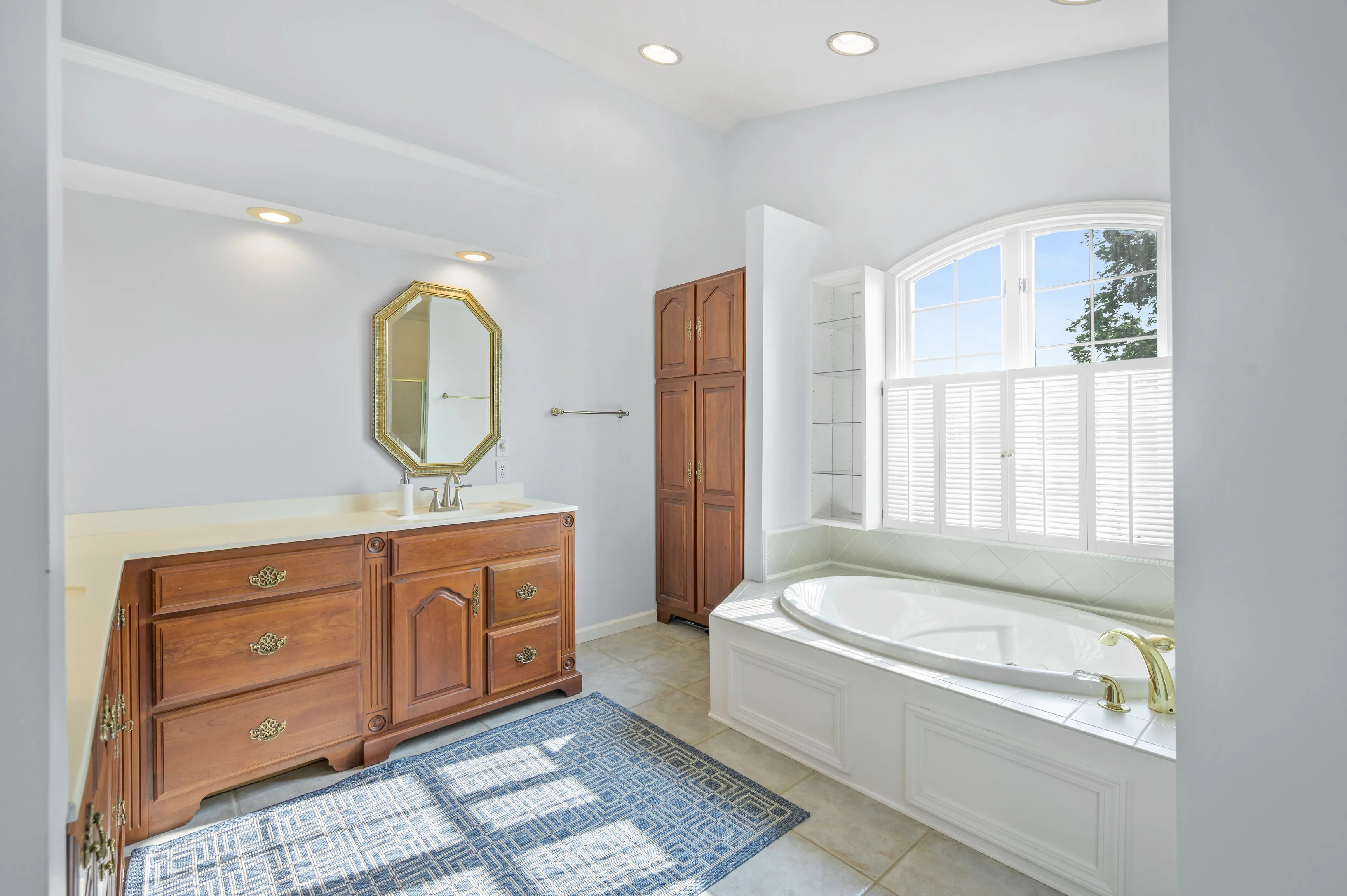 Bright modern bathroom with a large bathtub, wooden vanity cabinet, octagonal mirror, and a window with natural light.