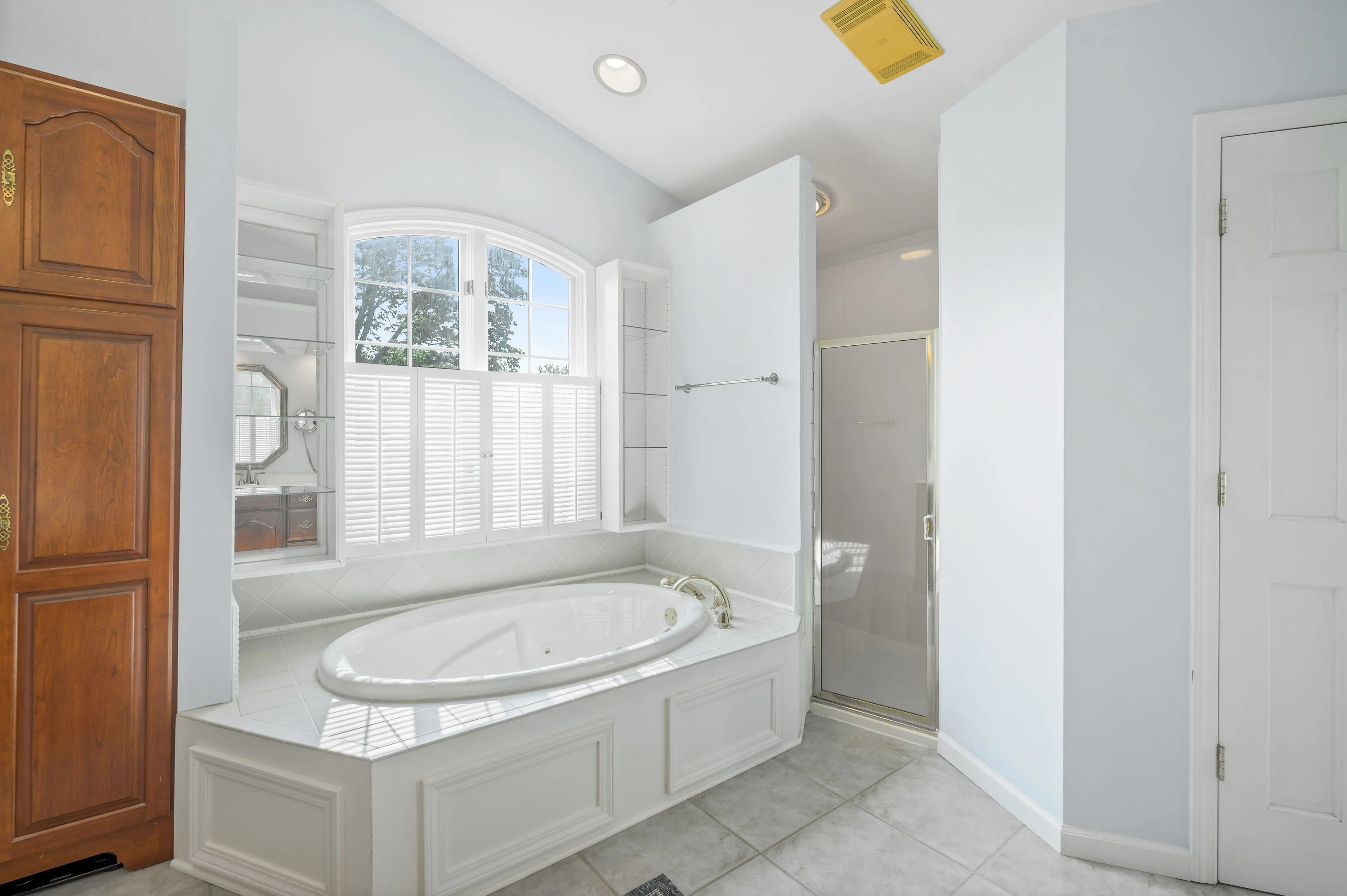 Bright bathroom interior with a corner jacuzzi tub, separate shower stall, wooden cabinet, and large window with blinds.
