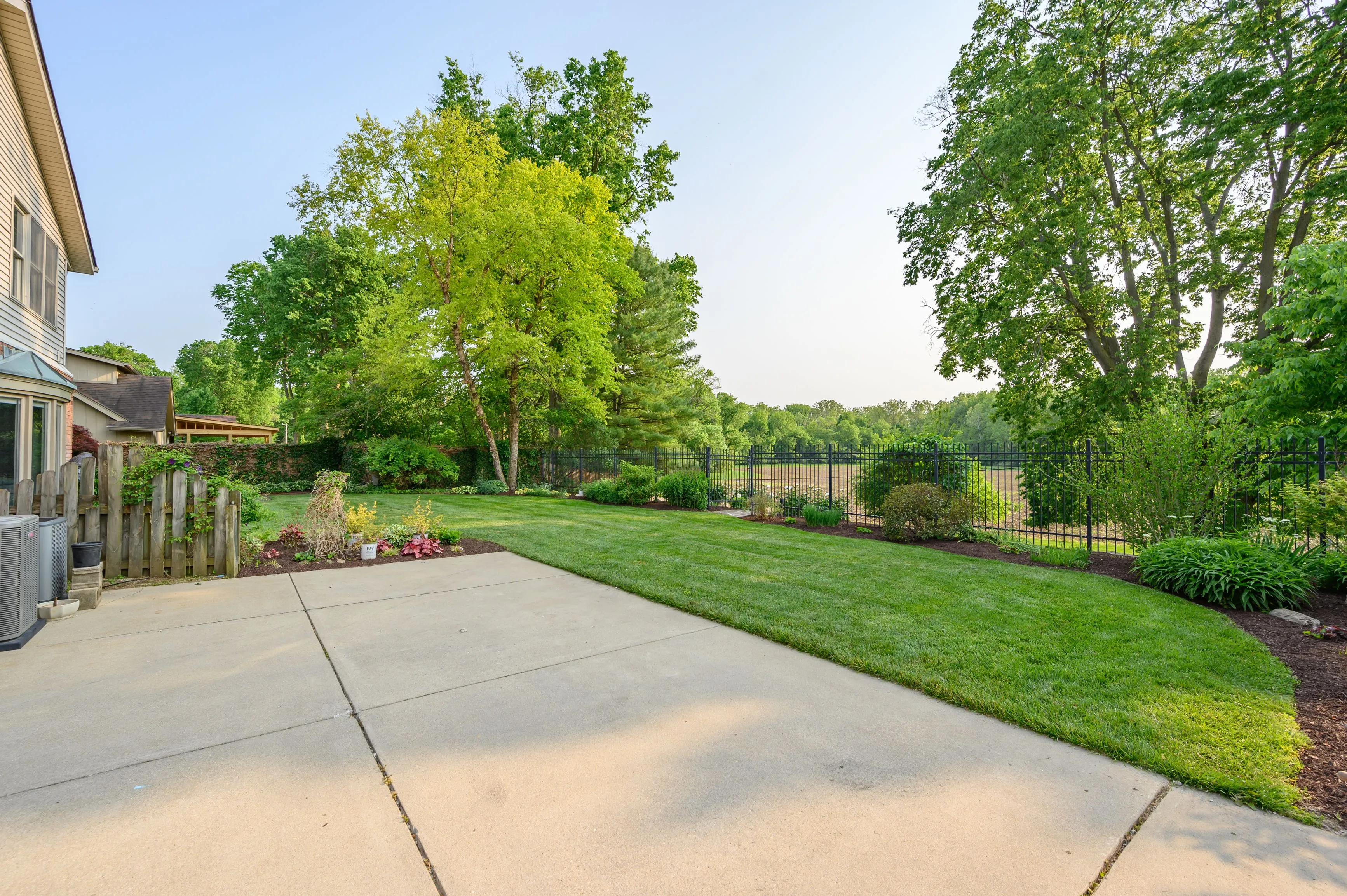  A concrete driveway leading to a residential backyard with lush green trees and lawn on a sunny day.