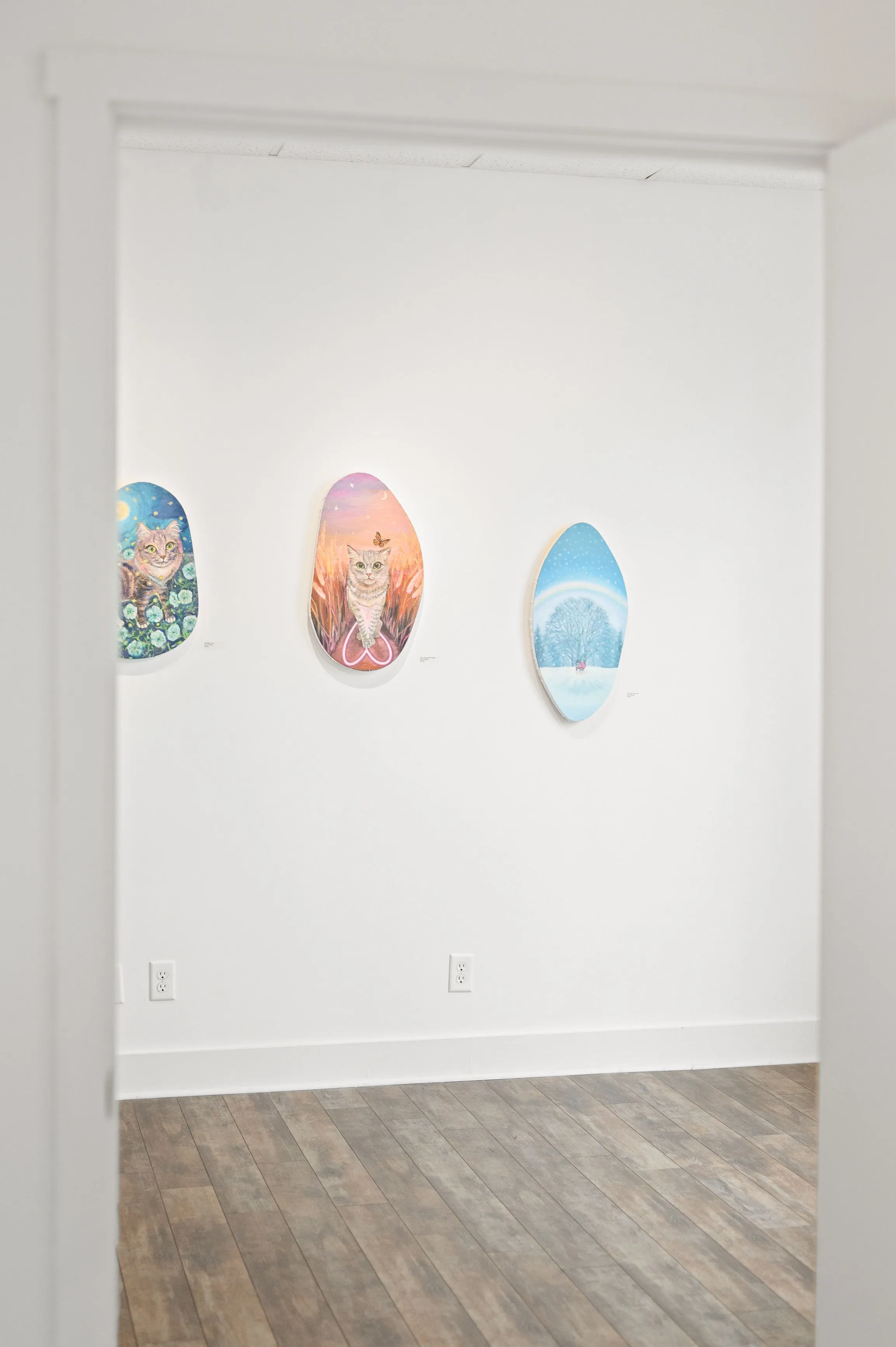 Three small, colorful abstract paintings displayed on a white wall in a bright room with wooden flooring.