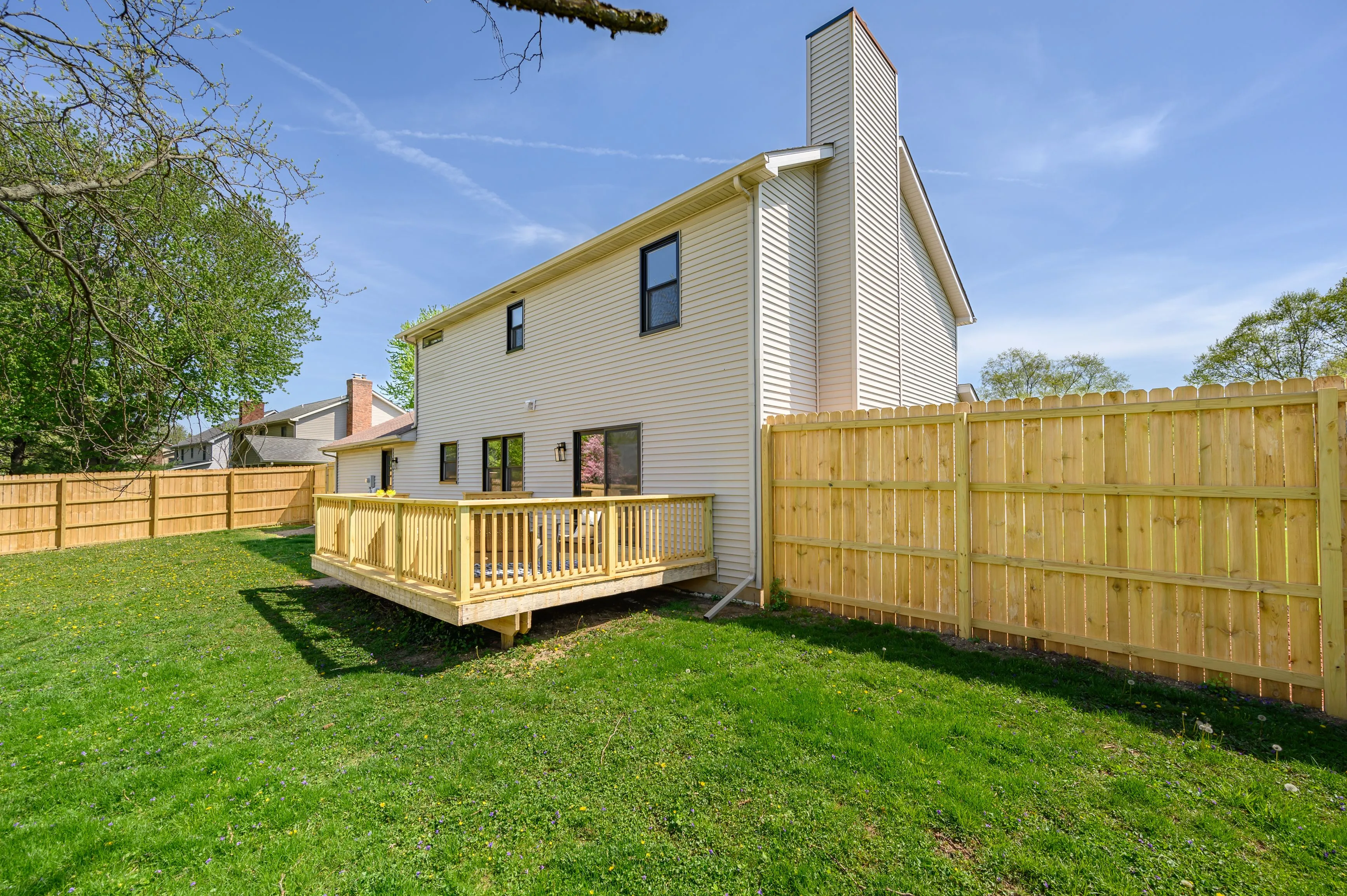Two-story house with a wooden deck and fenced backyard on a sunny day.