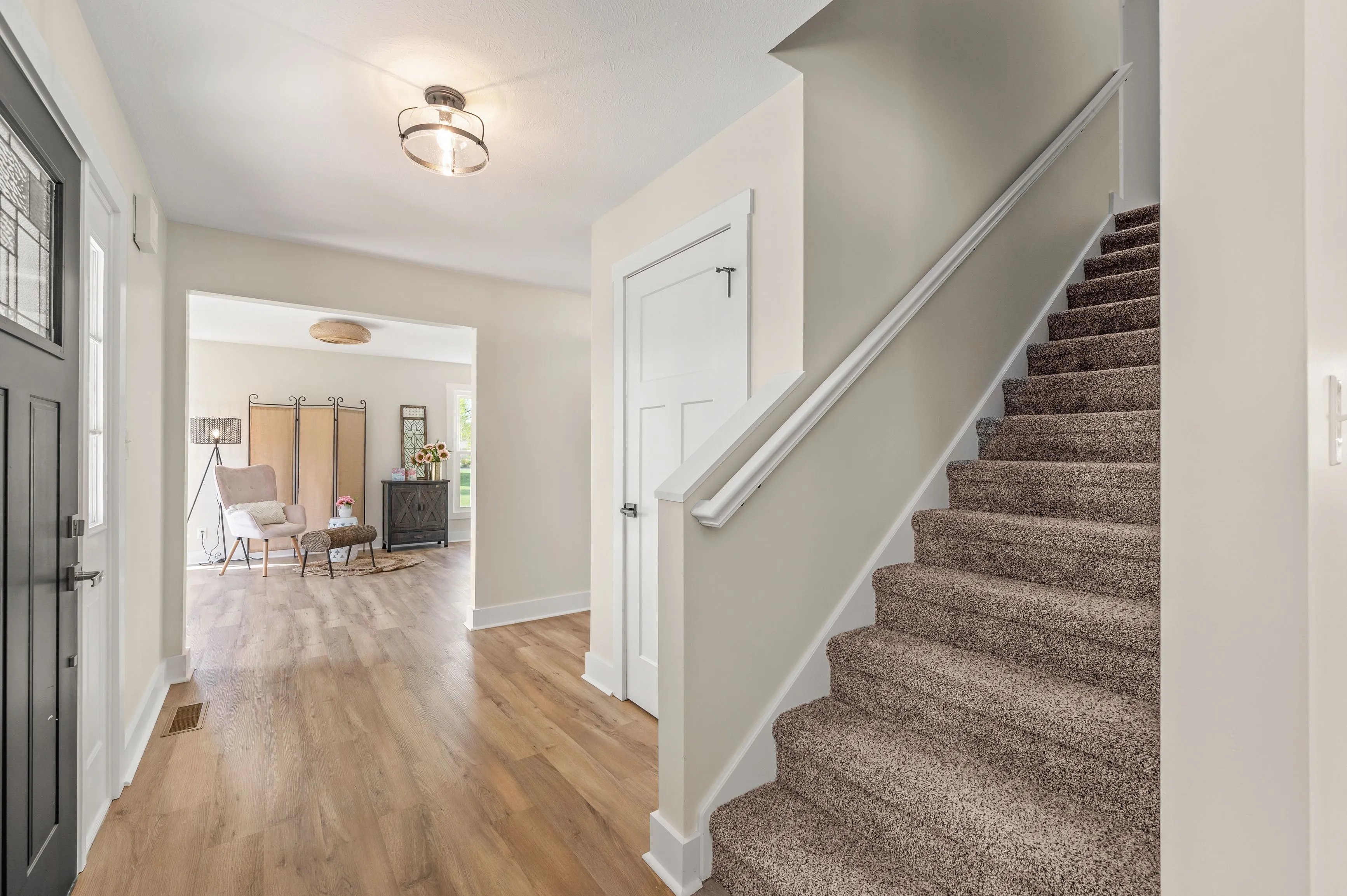 Interior view of a modern home entryway with hardwood floors leading to a staircase with carpeted steps and a well-lit, inviting living area in the background.