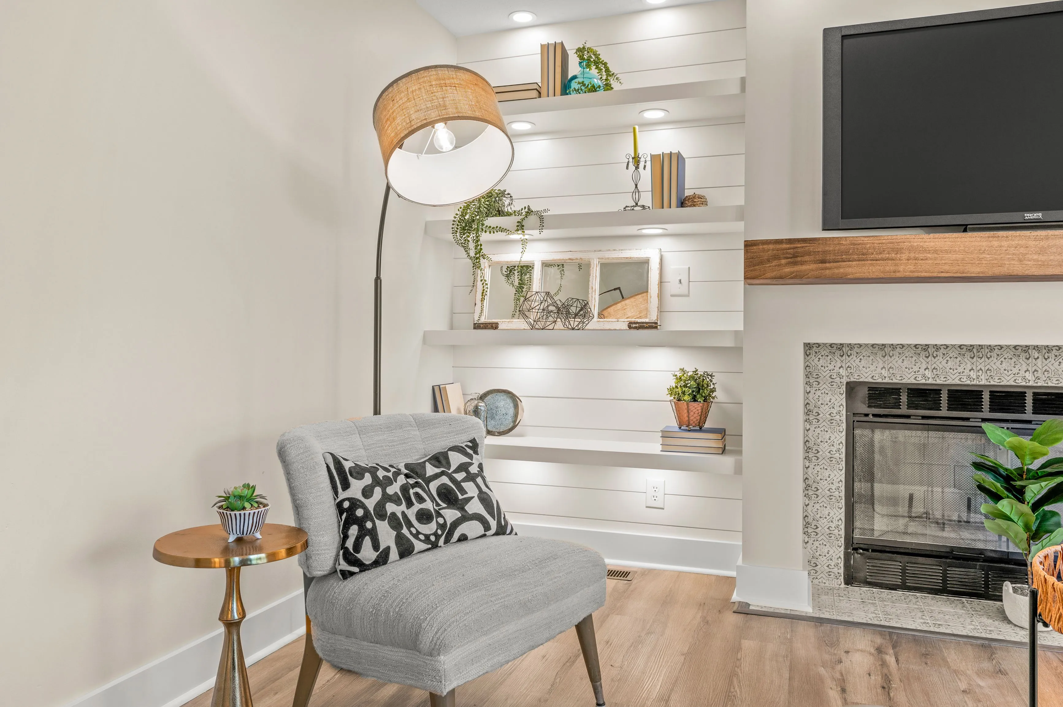 Cozy living room corner with a gray armchair, patterned cushion, floor lamp, side table, floating shelves, television, and a fireplace with green plants.