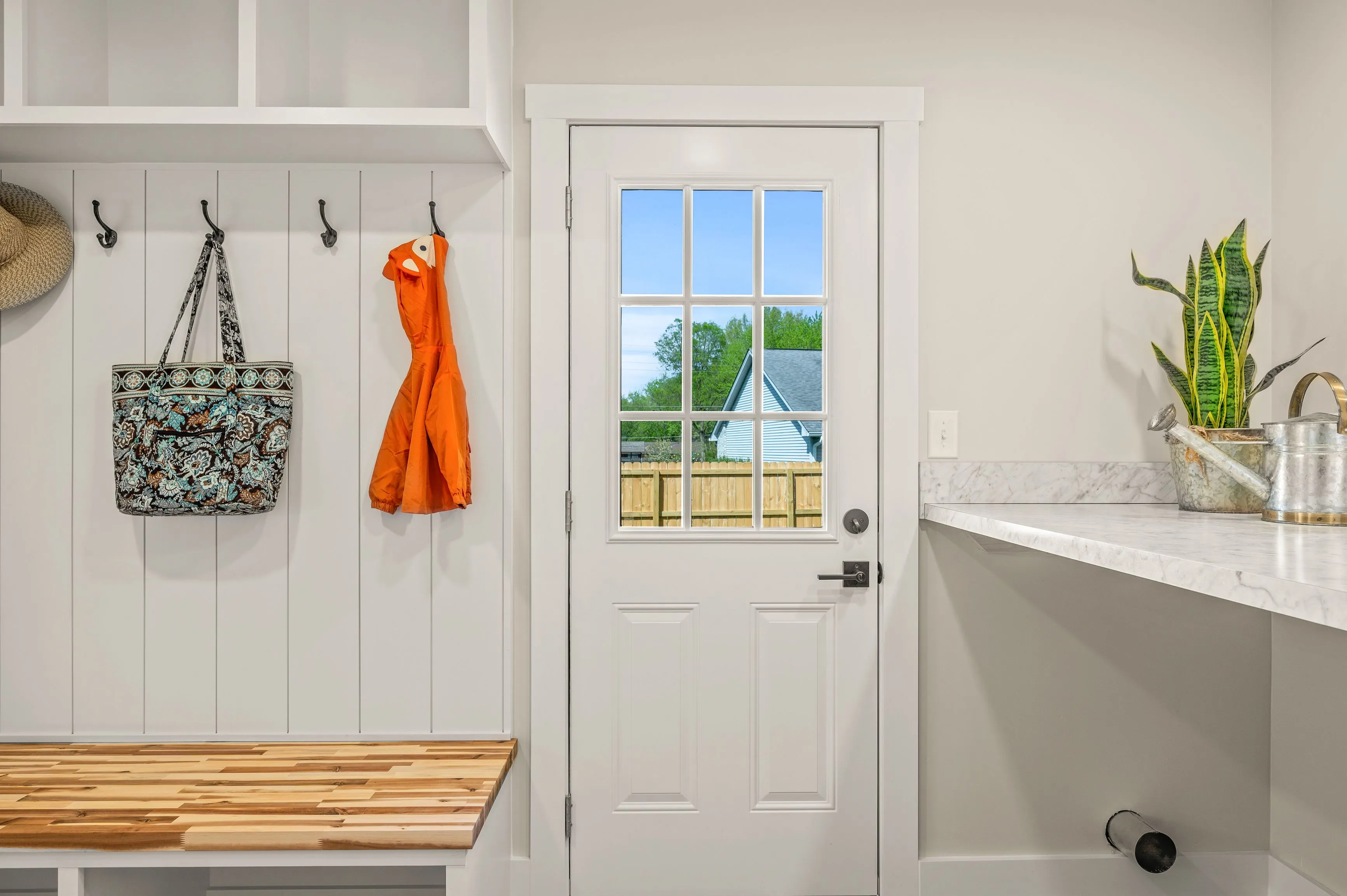 Bright, modern mudroom with white walls, a bench seat, hooks with hanging bags, and a back door with a window showing outdoor greenery.