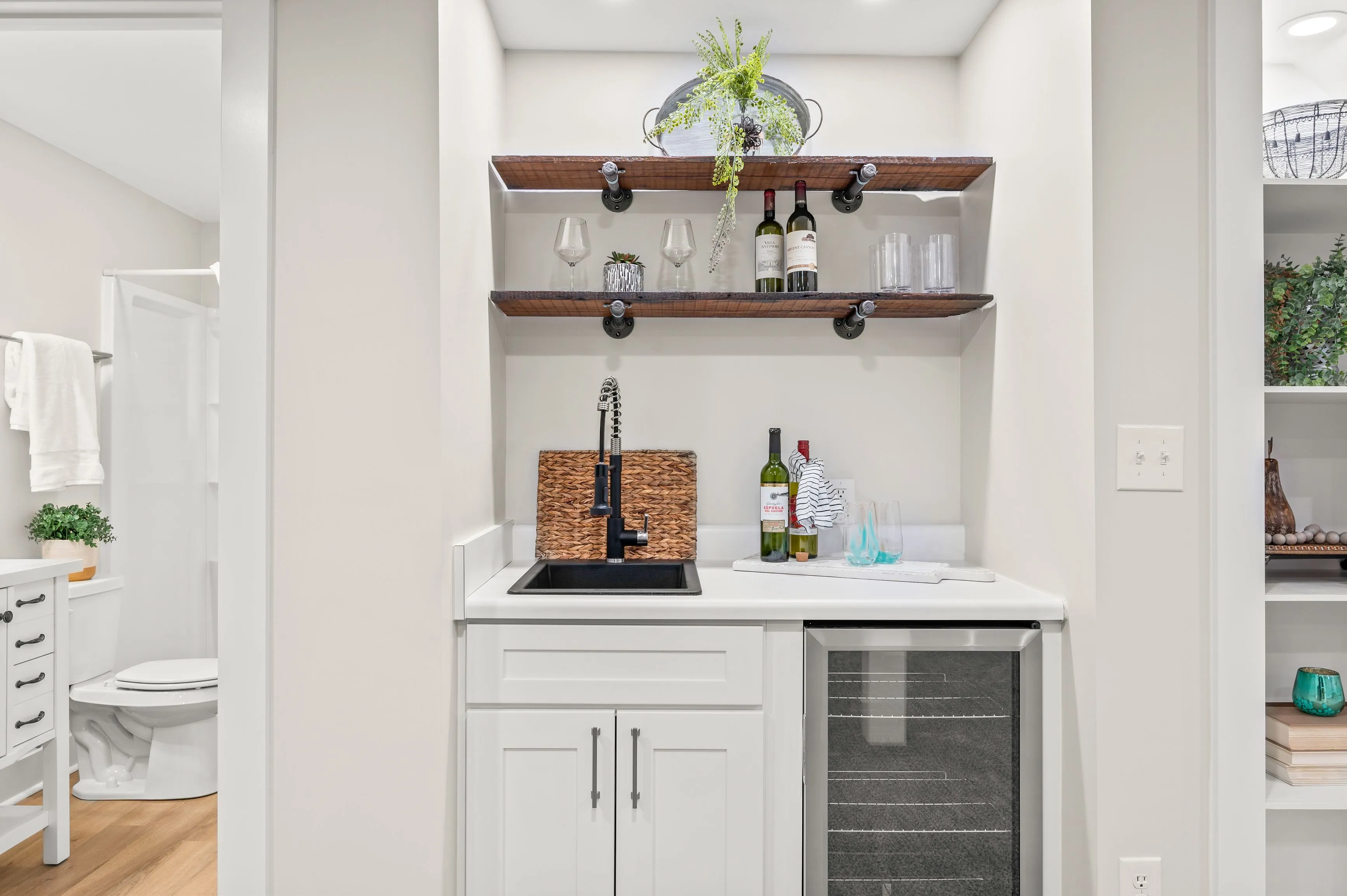 Modern kitchenette with white cabinets, stainless steel mini-fridge, and open wooden shelves with wine glasses and decor.