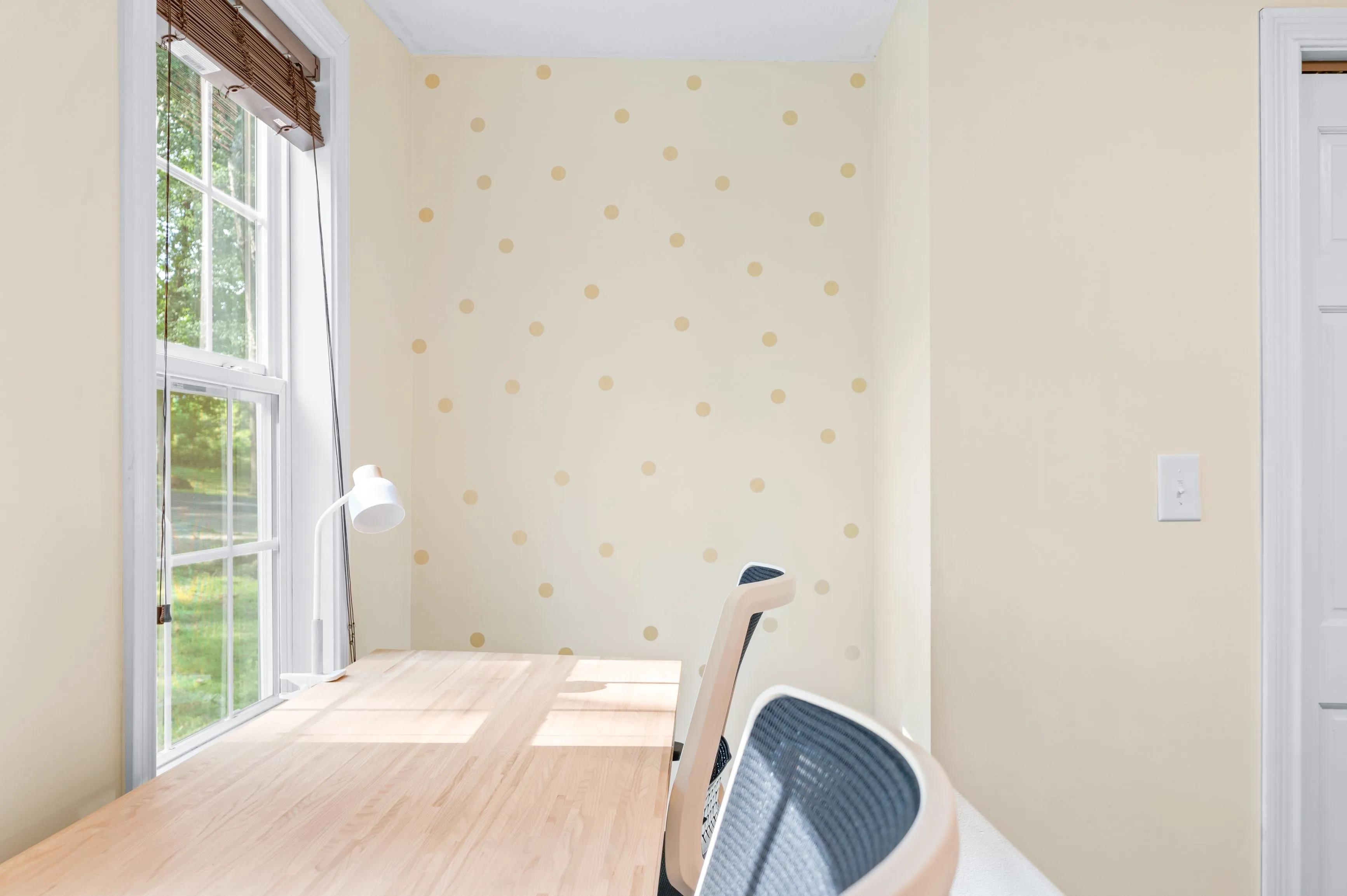 Bright, empty room with floral wallpaper, hardwood floor, and a window allowing natural light in, with a glimpse of a chair and a laundry basket in the foreground.