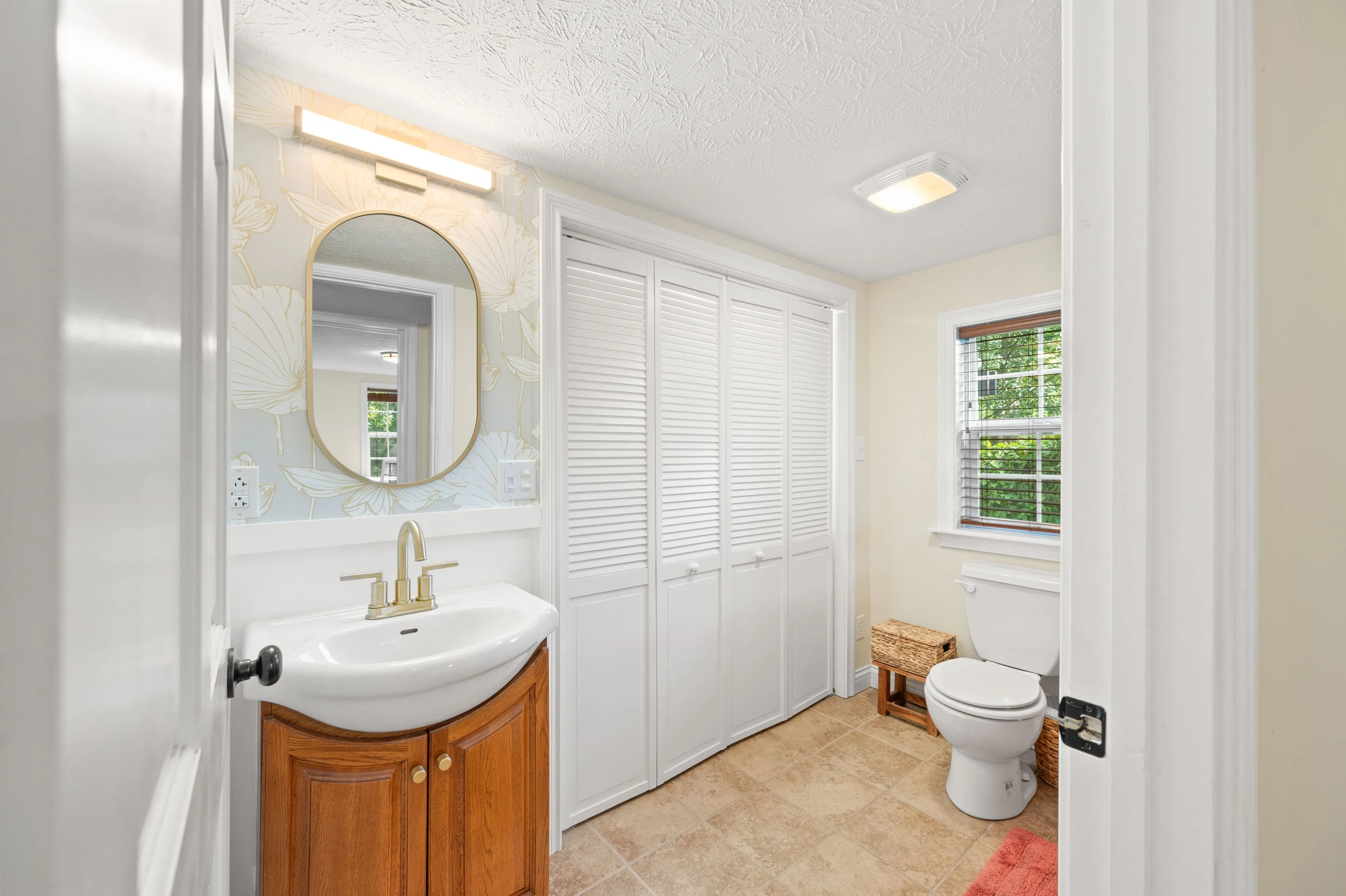 Bright, compact bathroom with white walls, a vanity with a wood cabinet, oval mirror, bathtub with a white shower curtain, and a toilet next to a window with a view of greenery.
