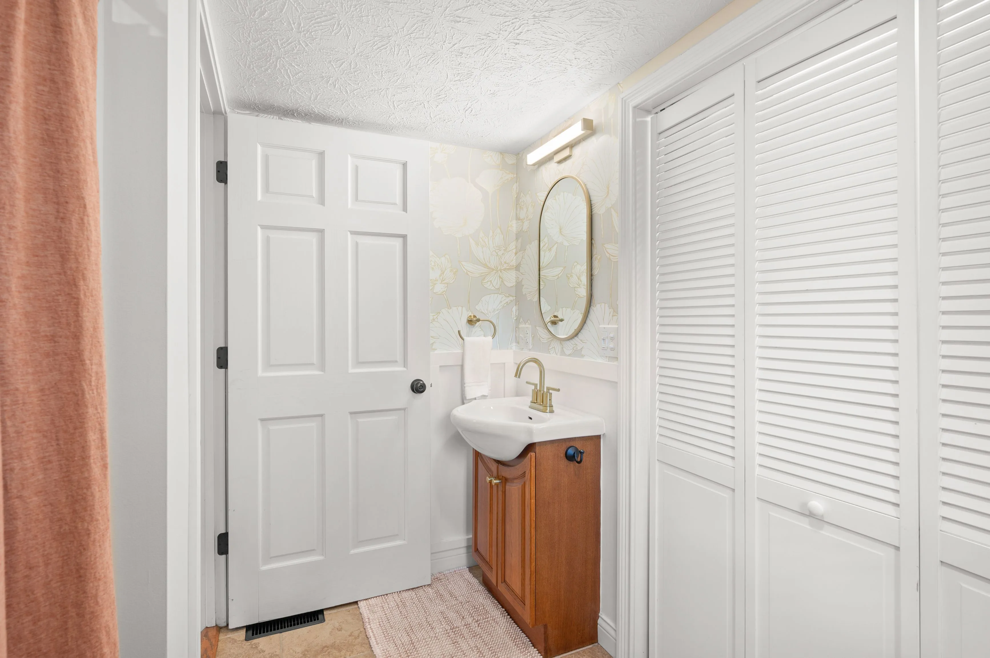 A small, narrow bathroom with white walls, a single vanity with wooden cabinets, a white sink, a mirror above the sink, and louvered doors on the right.