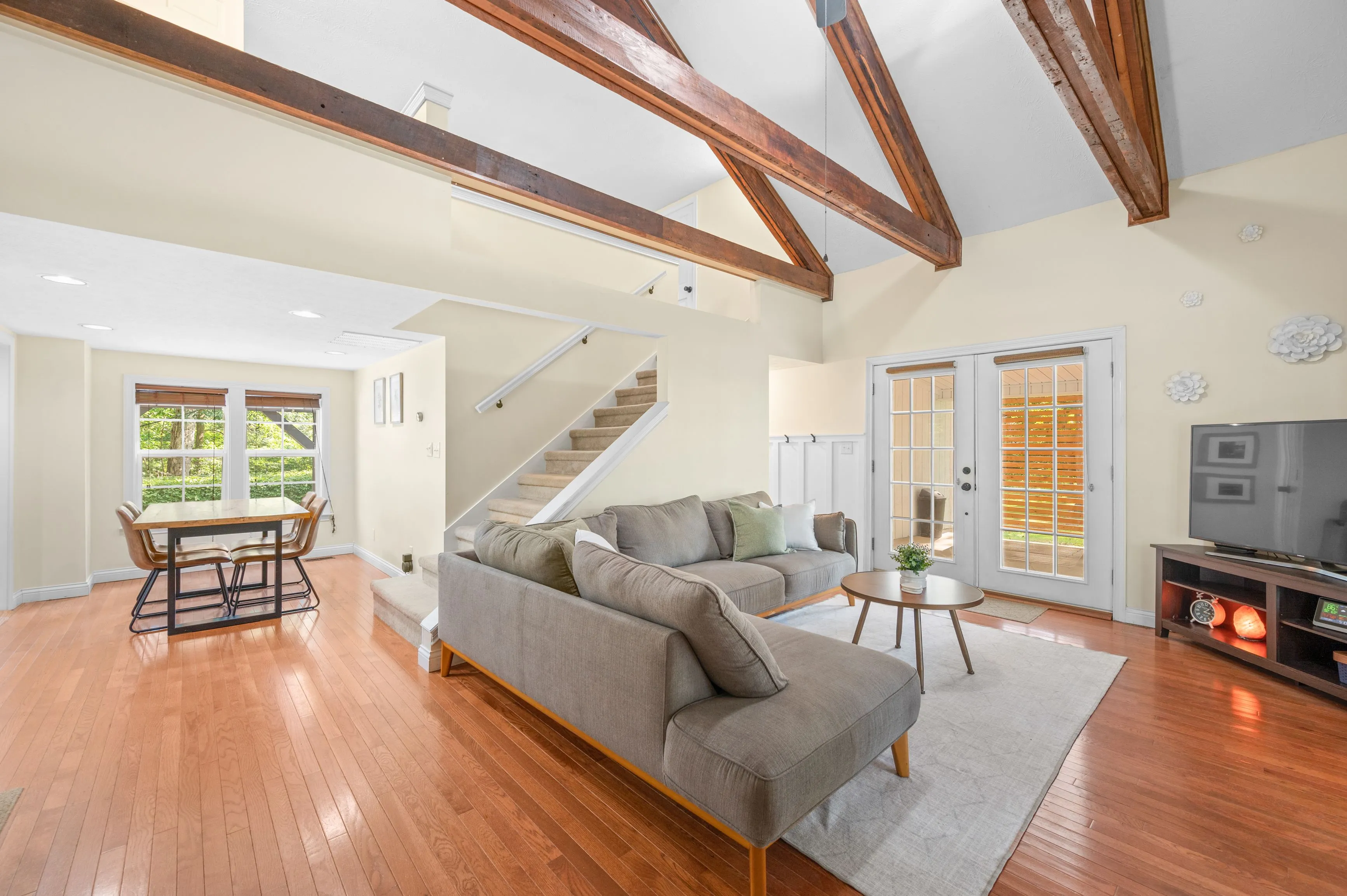 Bright living room interior with hardwood floors, a sectional sofa, a fireplace, exposed wooden beams on the ceiling, and stairs leading to the upper level.