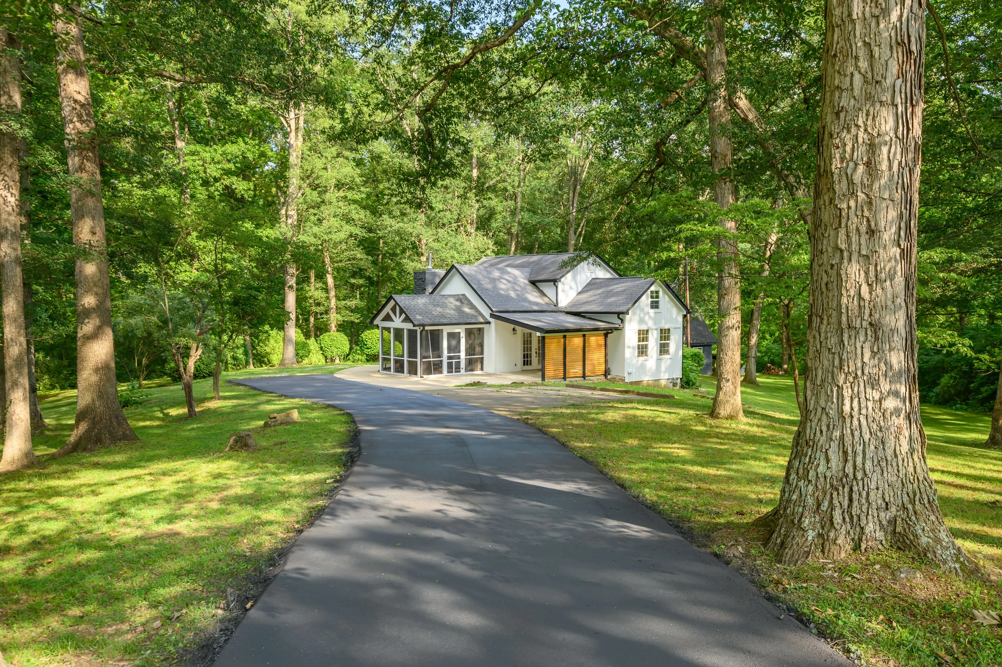 A serene suburban house with a paved driveway, surrounded by lush green trees under a clear sky.
