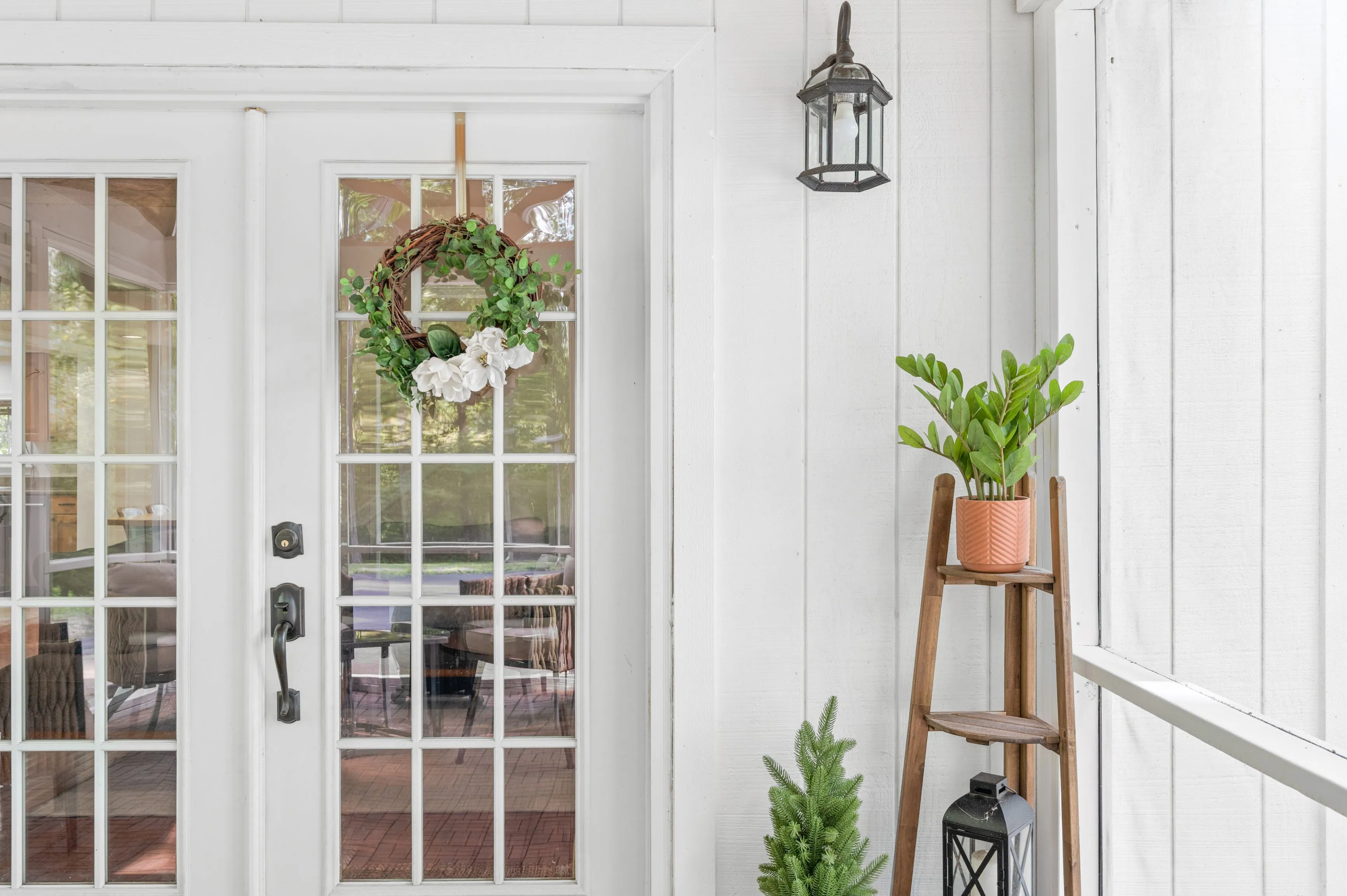 White entrance door with a wreath, glass panels, and a plant on a stool beside a hanging lantern.