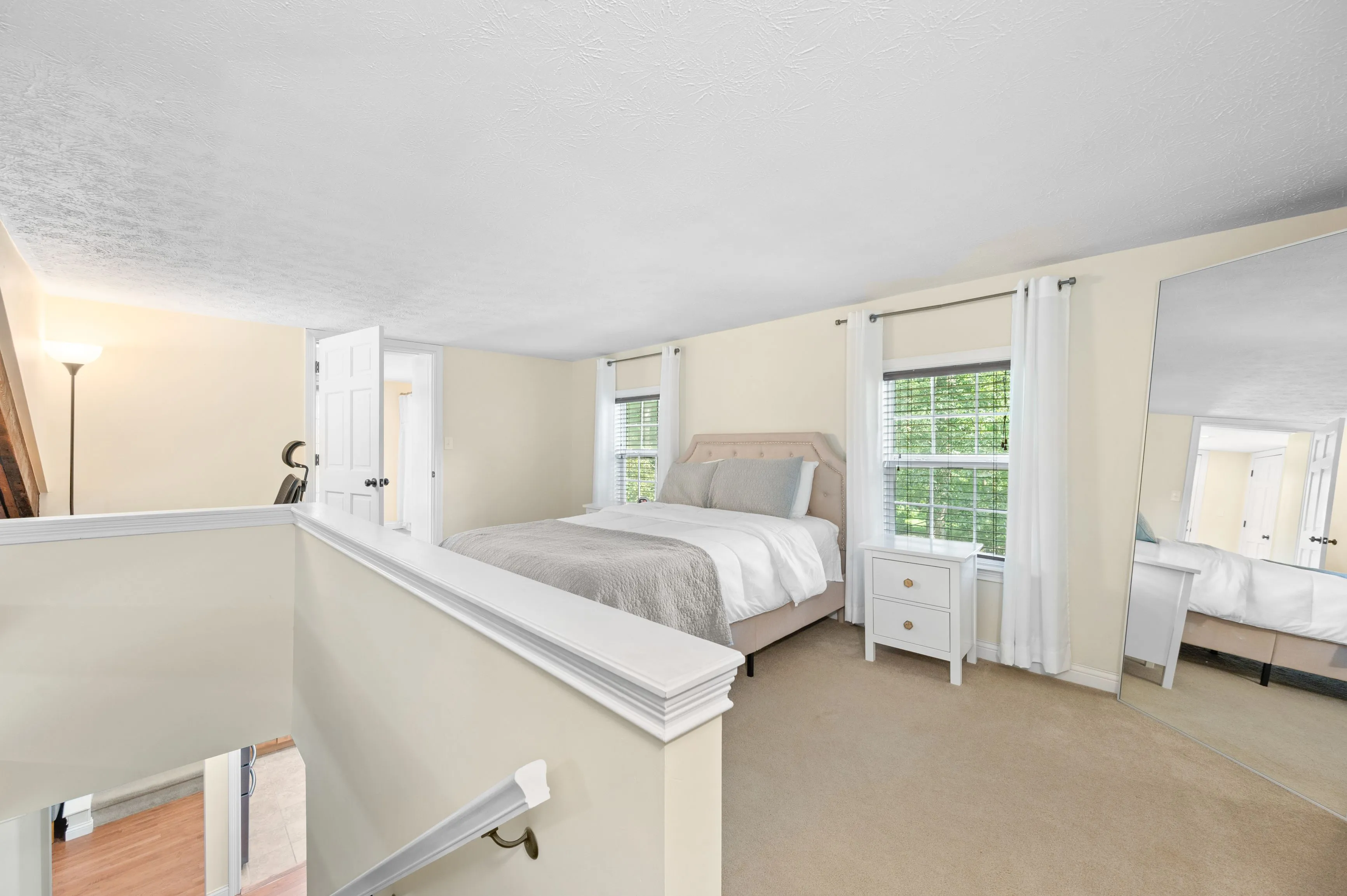 Bright and airy bedroom interior with queen-sized bed, white linens, double windows, and an open door leading to an adjoining room.