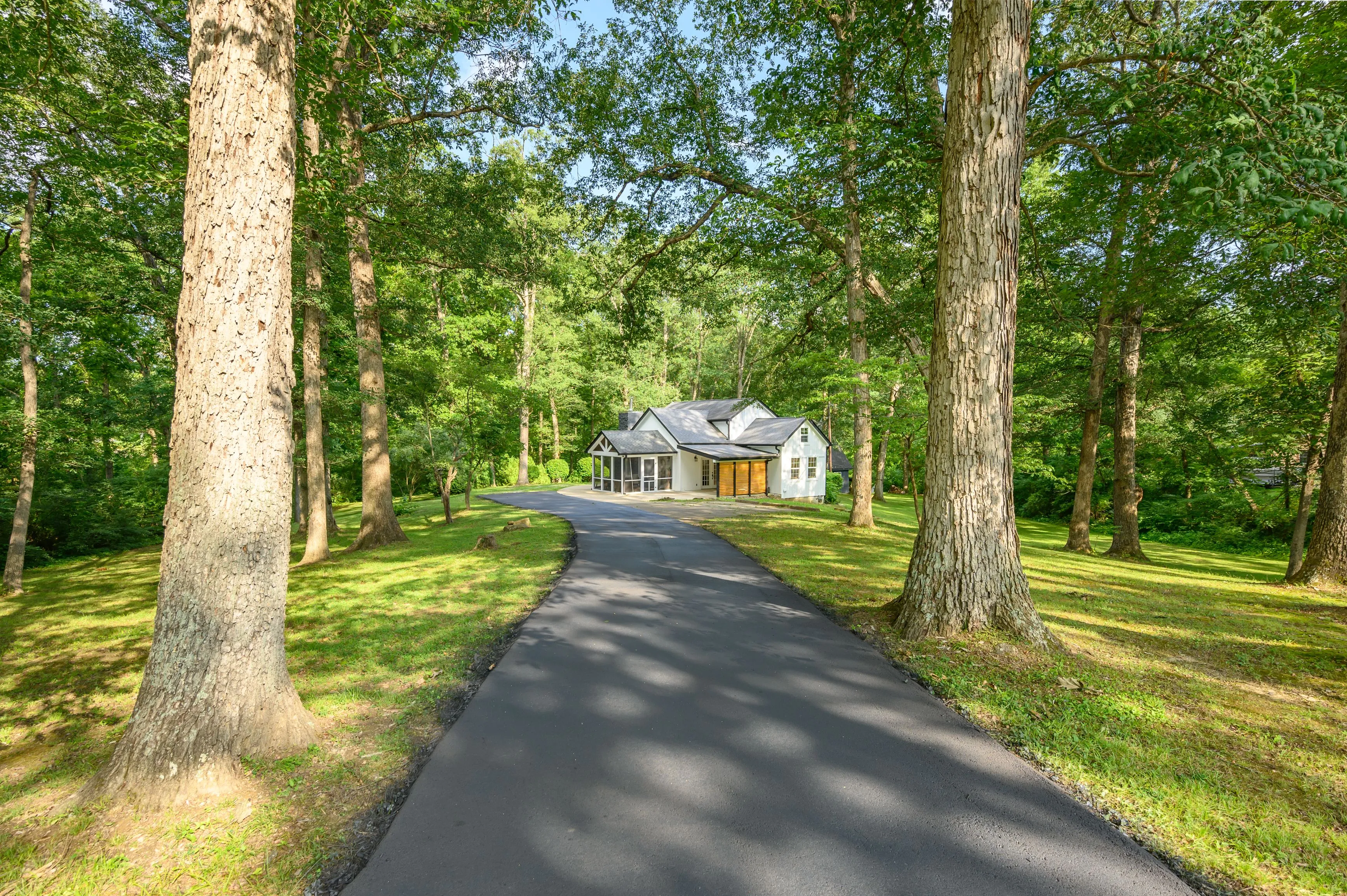 Paved driveway leading to a house surrounded by lush green trees on a sunny day.