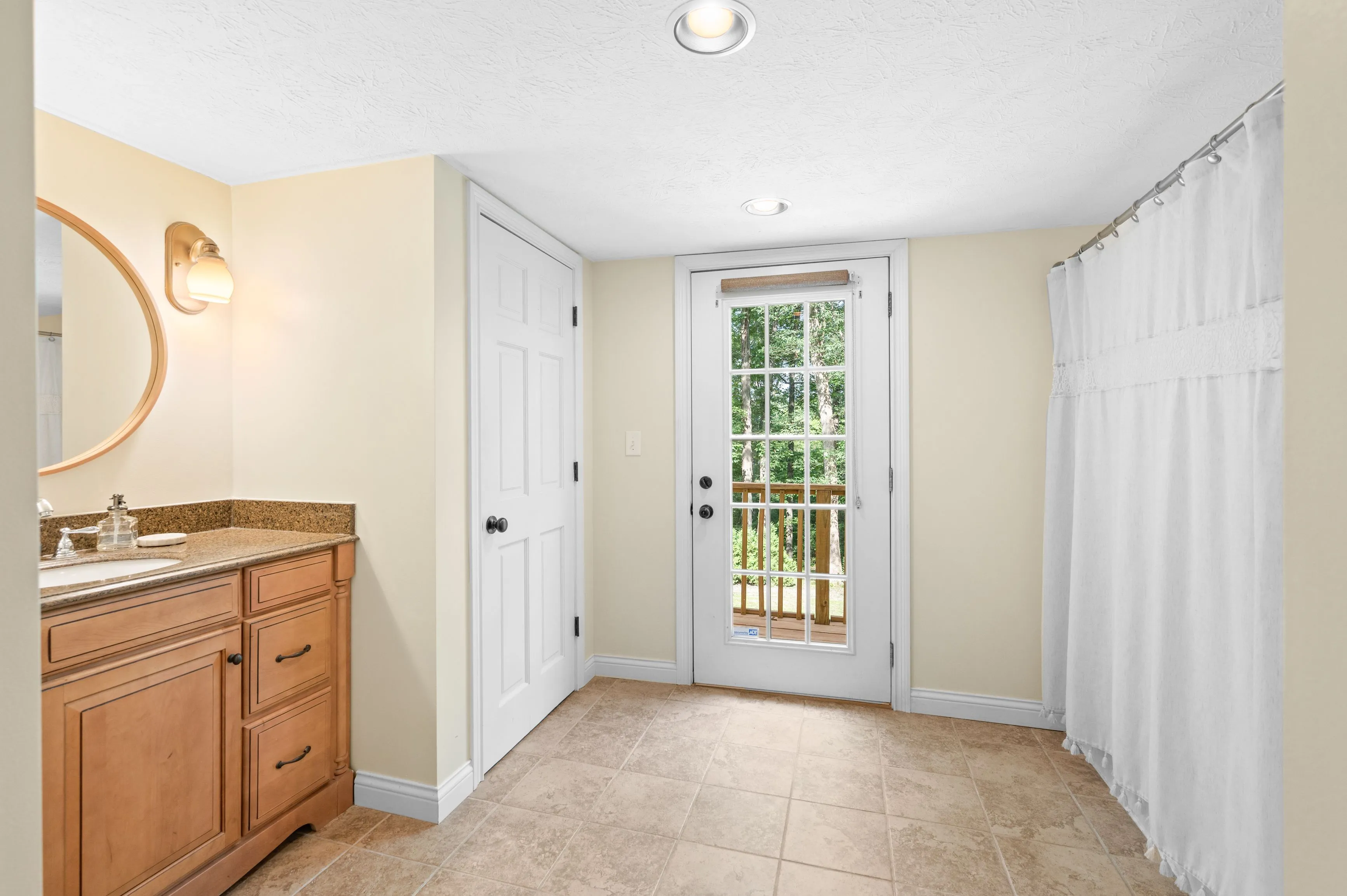 Bright, empty room with tiled floor, wooden vanity and sink on the left, open French doors leading to outside, and white curtains.