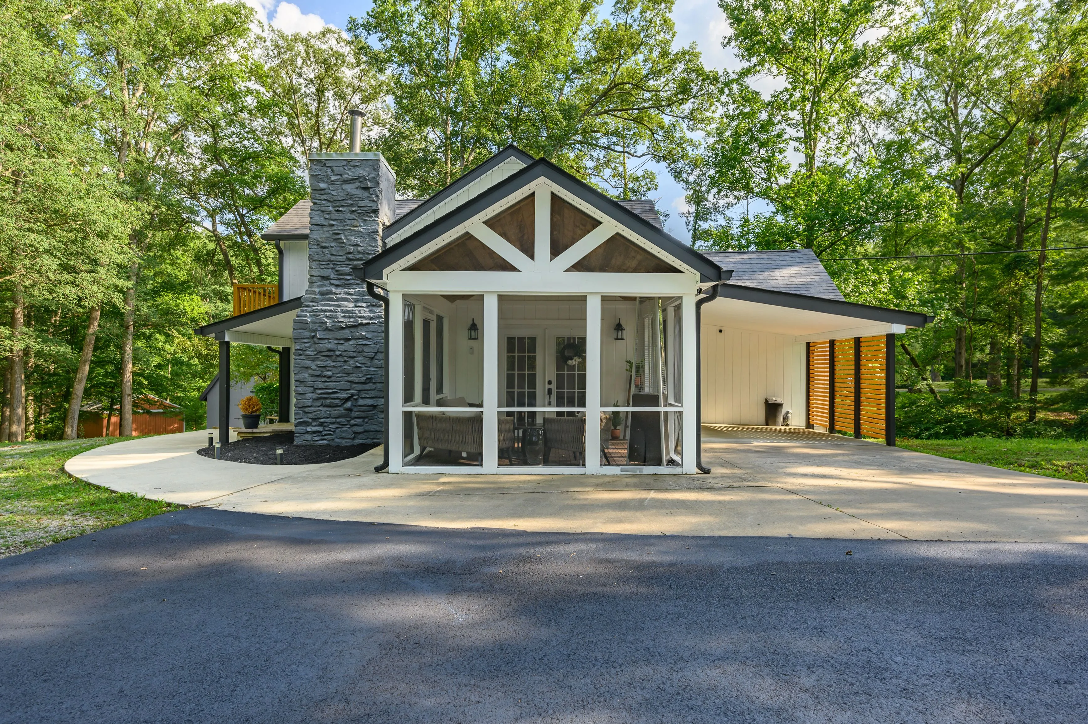 Modern single-family house with gable roof, stone chimney, and a paved driveway surrounded by trees.