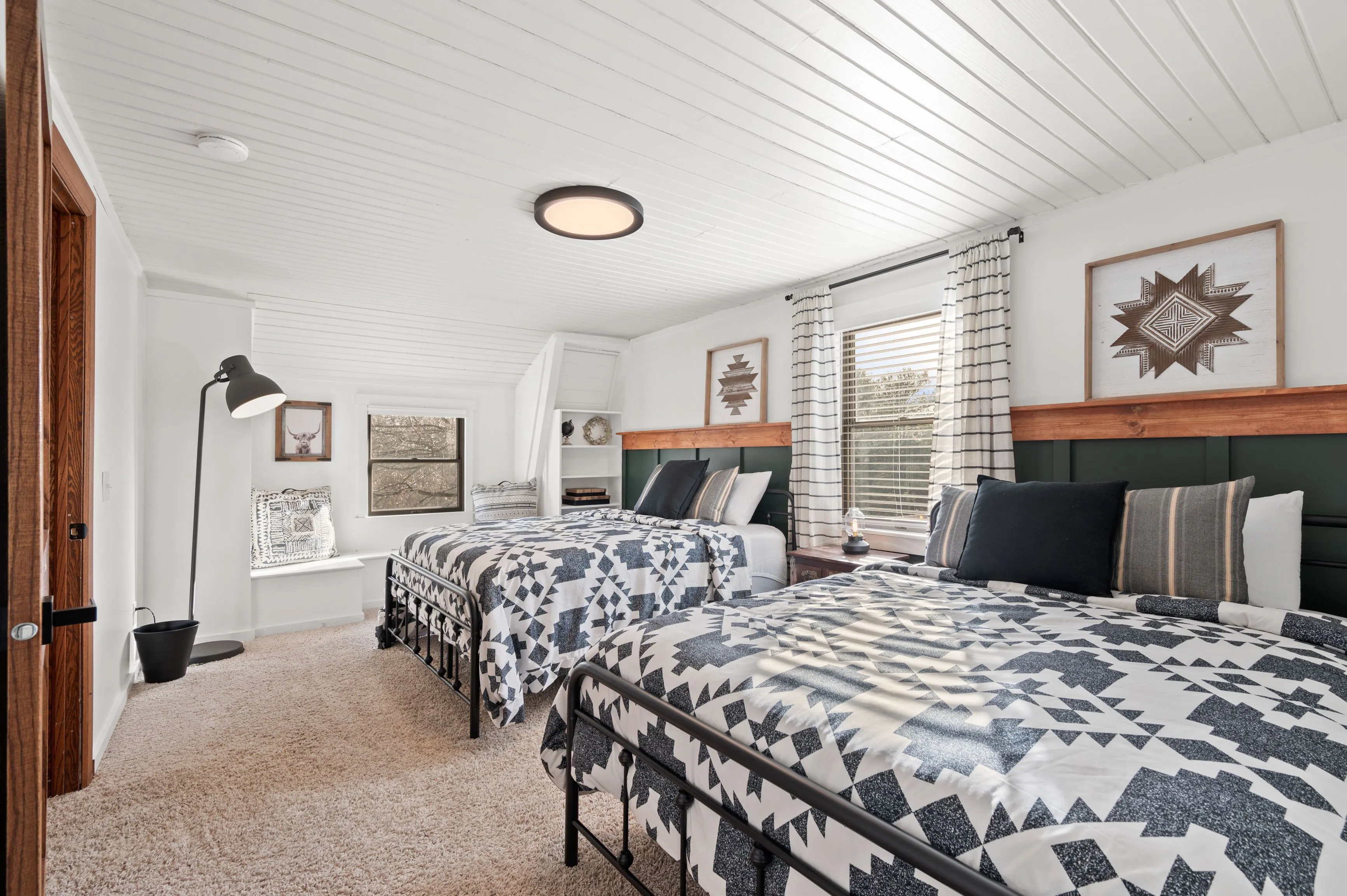 Bright and cozy bedroom with two single beds covered in matching quilts, white beadboard ceiling, window with blinds, and decorative wall art.