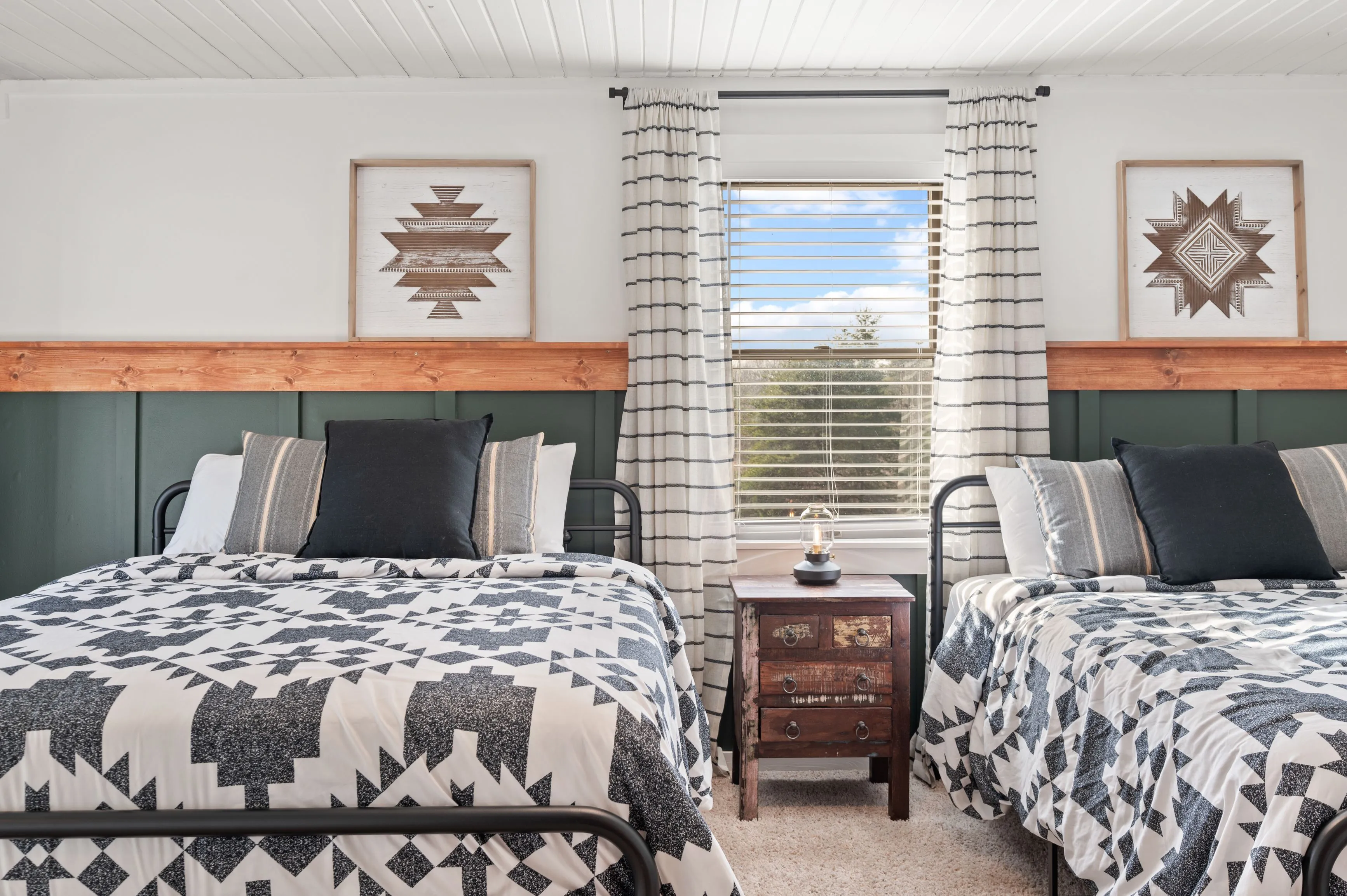 Cozy bedroom with two single beds with black and white geometric patterned bedding, rustic wooden headboards and decorative framed art above, dark green paneled walls, and a sunny window with striped curtains.