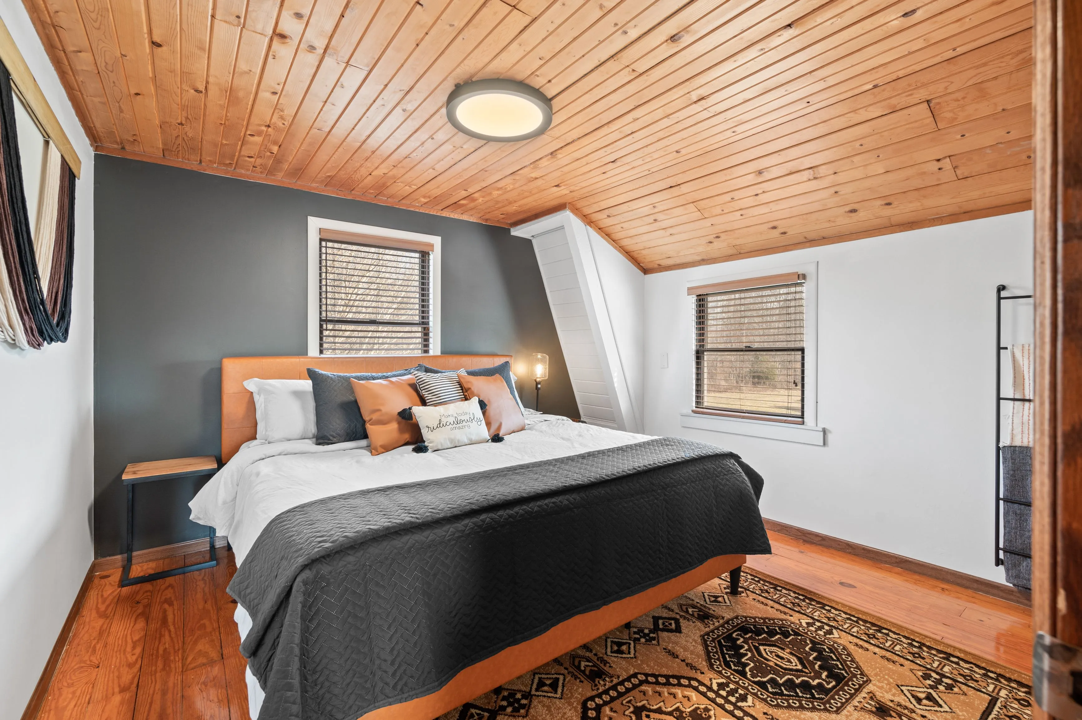 Cozy attic bedroom with slanted wooden ceiling, a large bed with decorative pillows, patterned rug, and a side table.