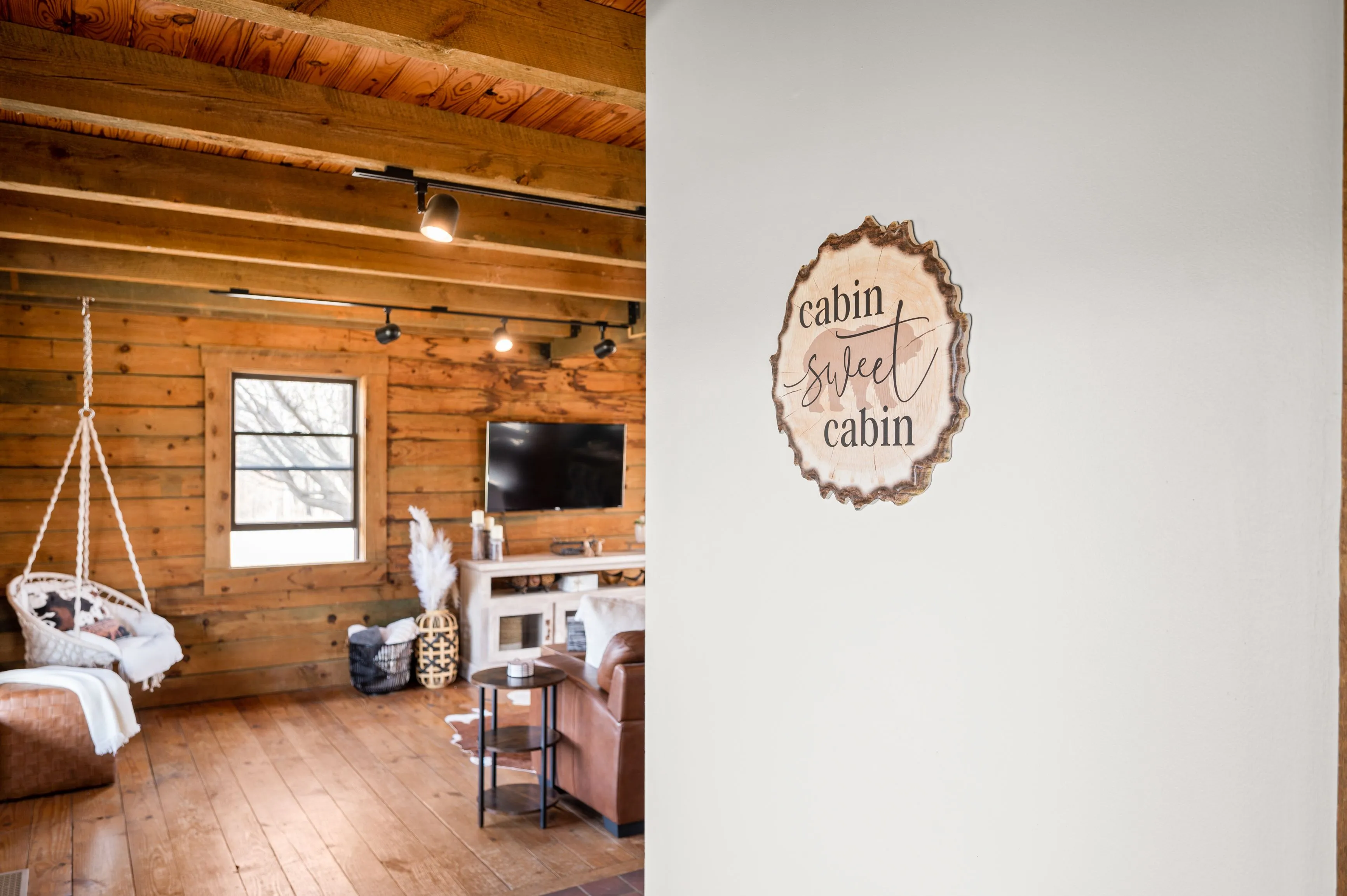 Interior of a rustic cabin living room with wooden walls and floors, featuring a swing chair, leather sofa, and a decorative sign that says "cabin sweet cabin".