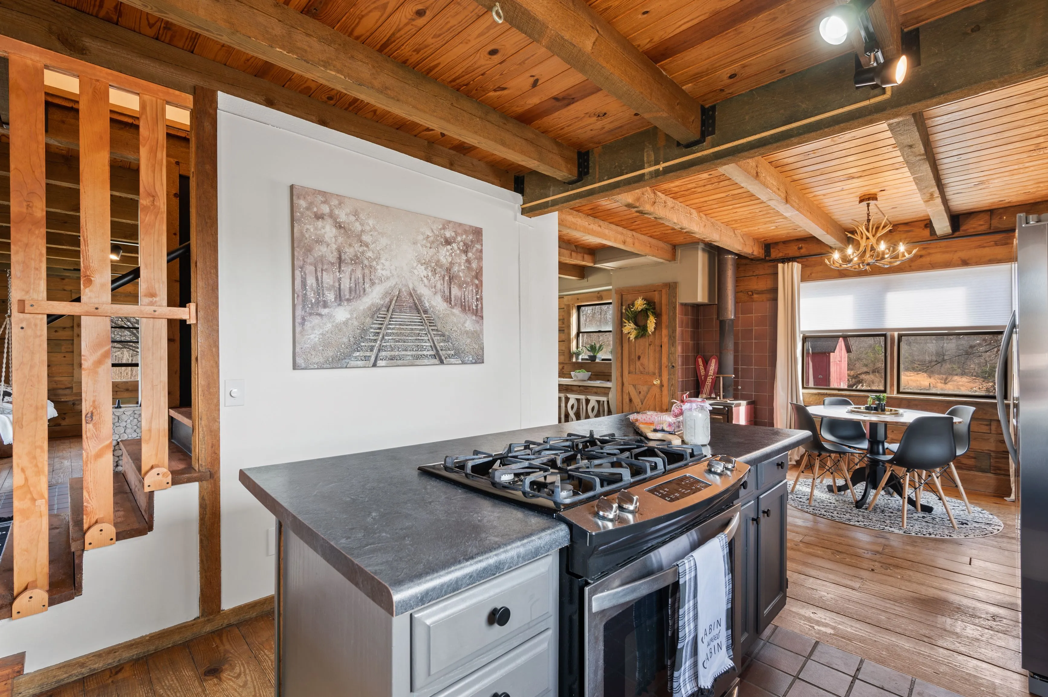 Rustic-style kitchen interior with exposed beams, hardwood floors, a gas stove, a painting of train tracks, and an adjoining dining area with a modern table set.