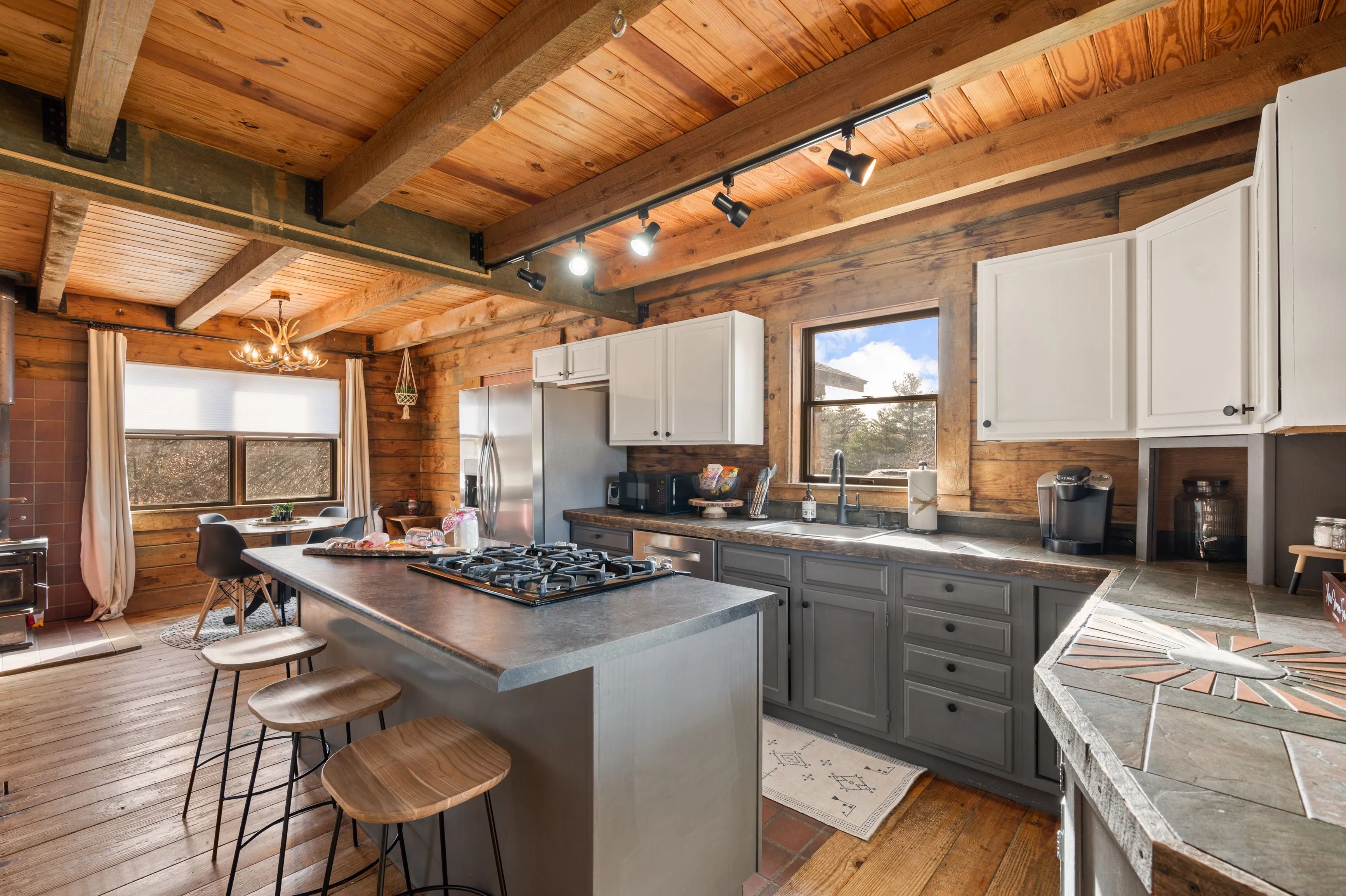 Rustic kitchen interior with wooden beams, hardwood floors, gray cabinetry, and a kitchen island with bar stools.