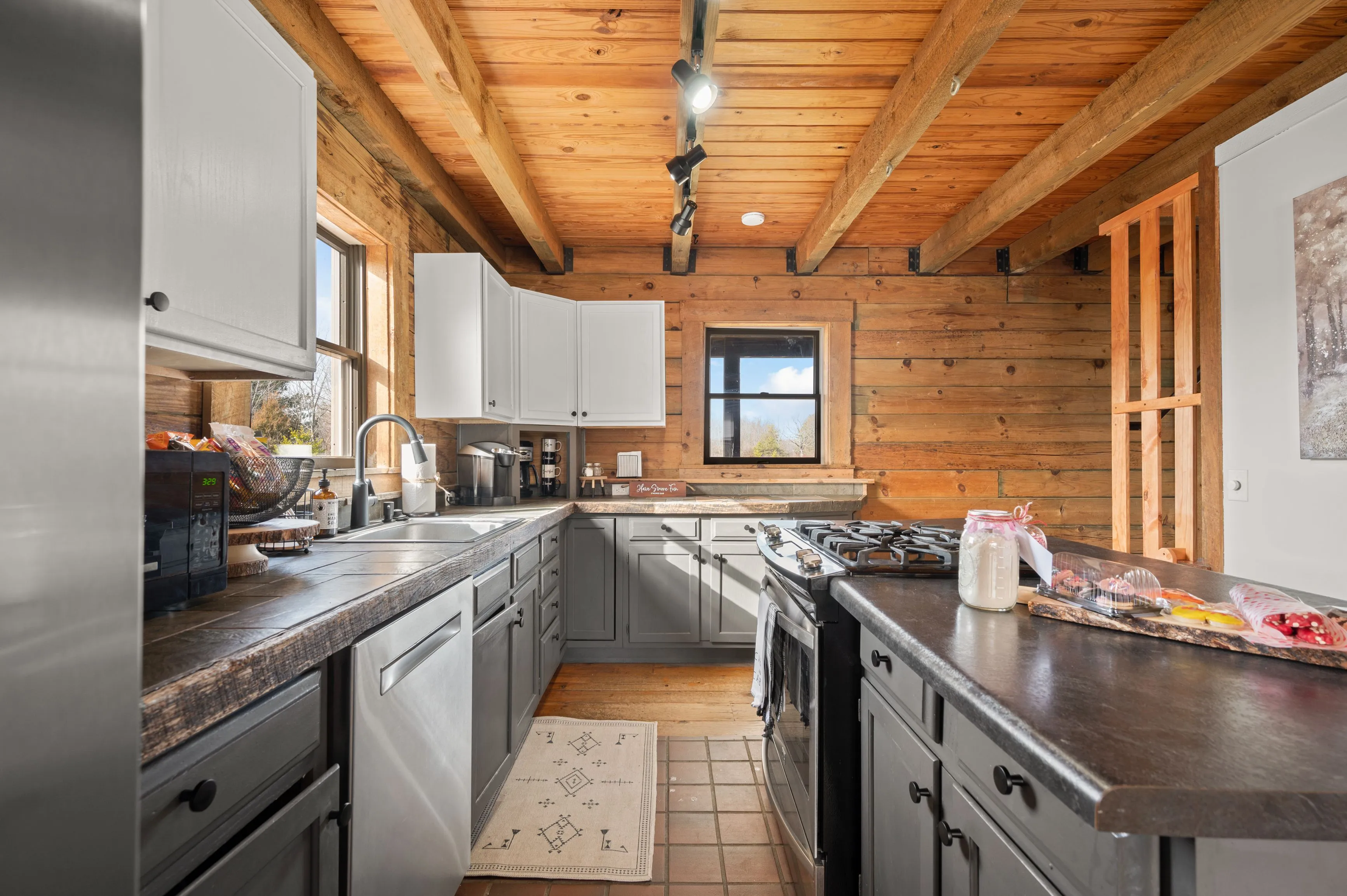 Rustic kitchen interior with wooden beams, log walls, white cabinets, stainless steel appliances, and black countertops illuminated by natural sunlight.
