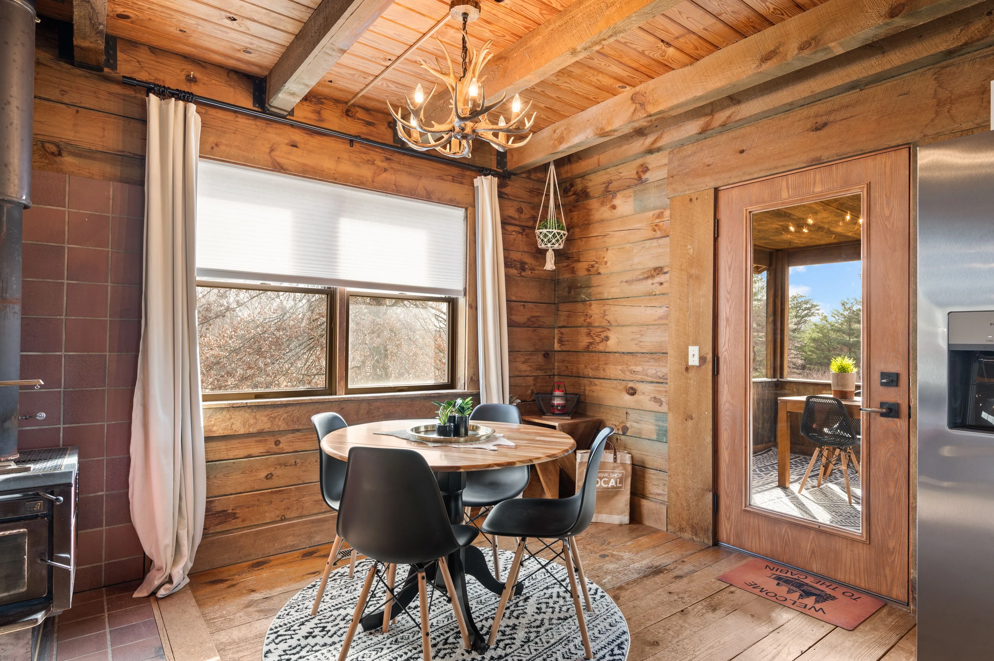 Cozy rustic dining area with a round wooden table, modern chairs, and a chandelier resembling antlers, inside a cabin with wood panel walls and ceiling, adjacent to a kitchen area with large windows providing natural light.