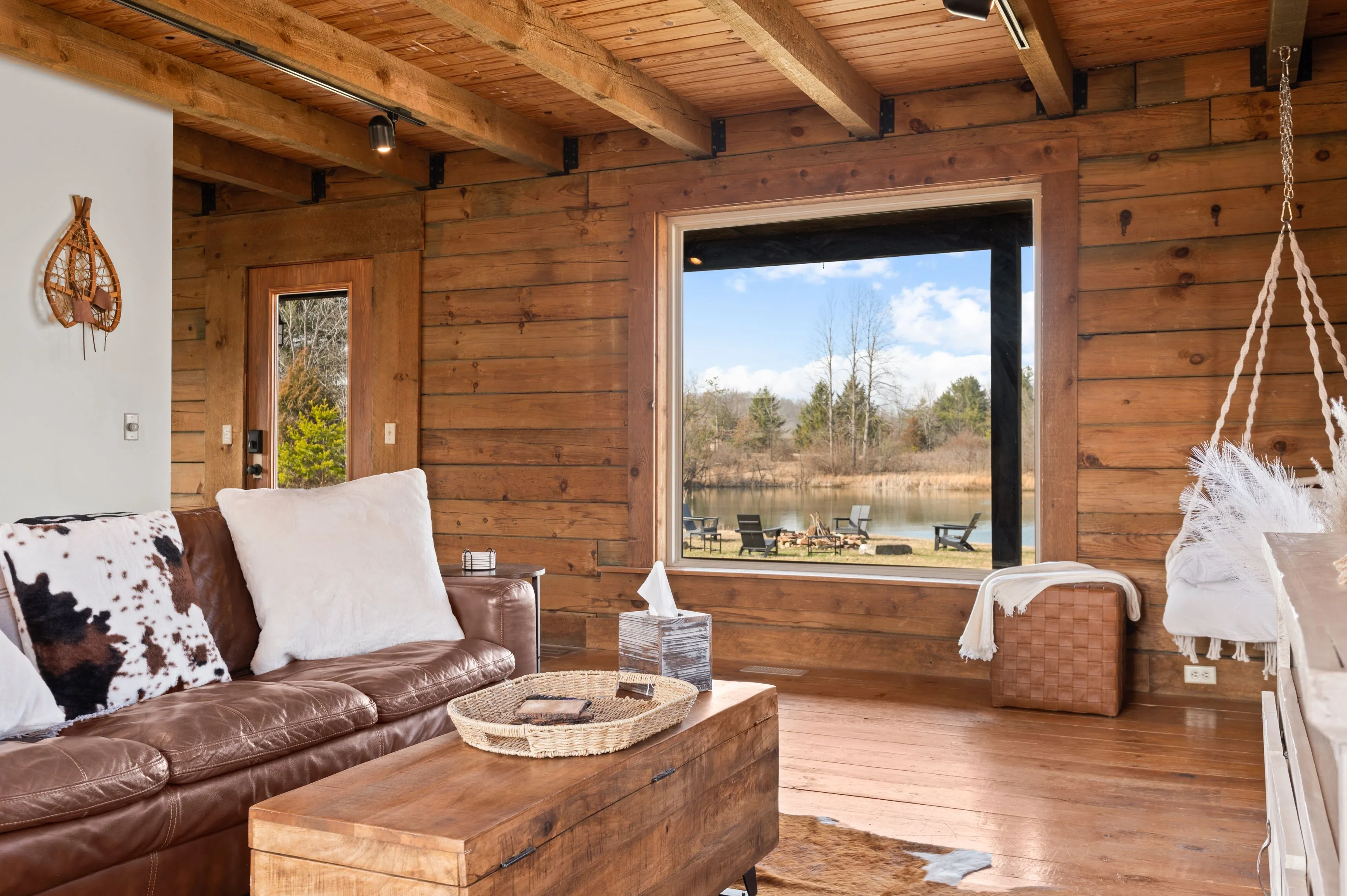 Cozy rustic living room with a leather sofa, wood coffee table, and large window overlooking a scenic outdoor area with a pond.