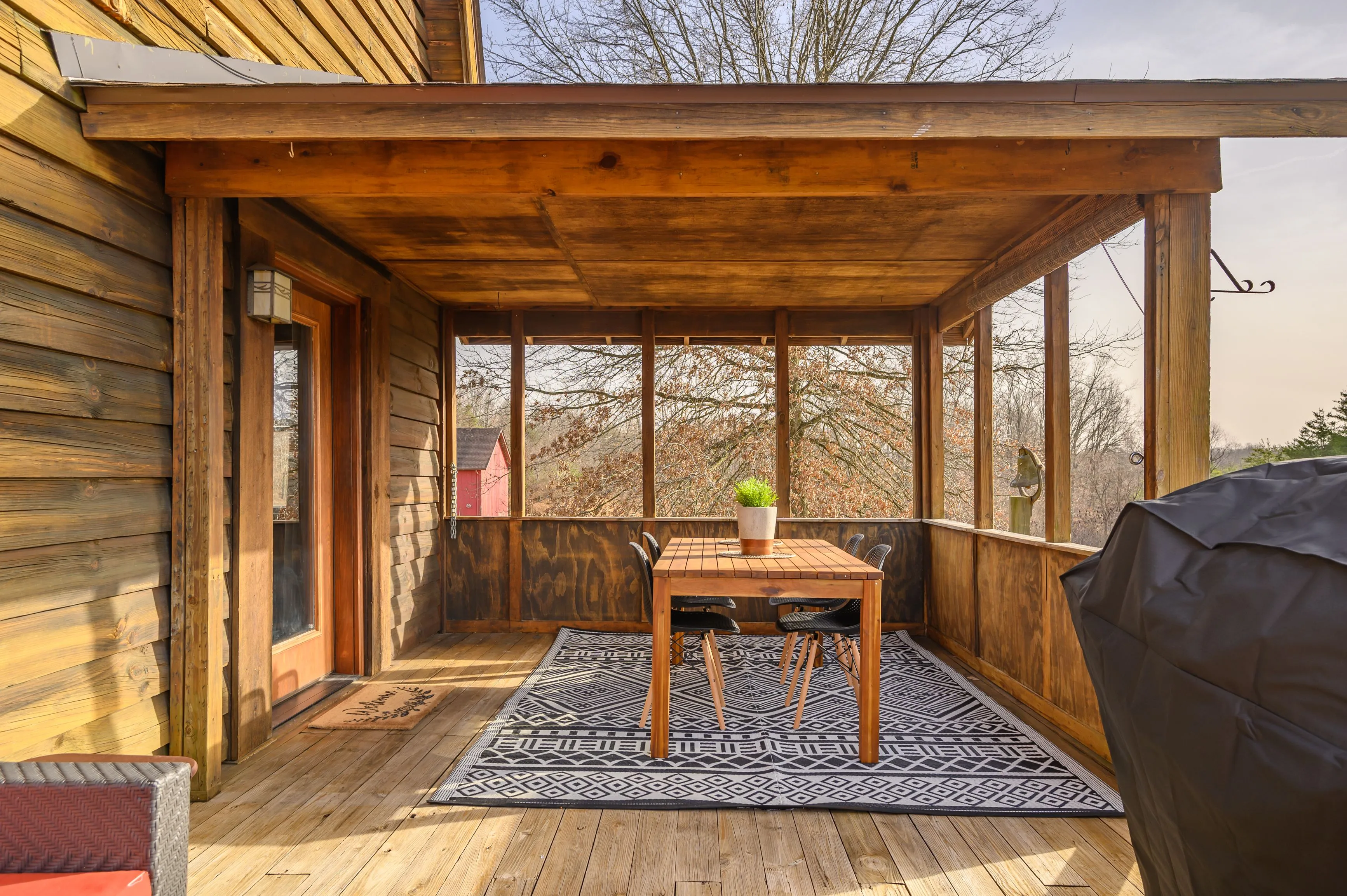 Cozy wooden screened porch with a dining table and chairs, patterned rug, and forest view.