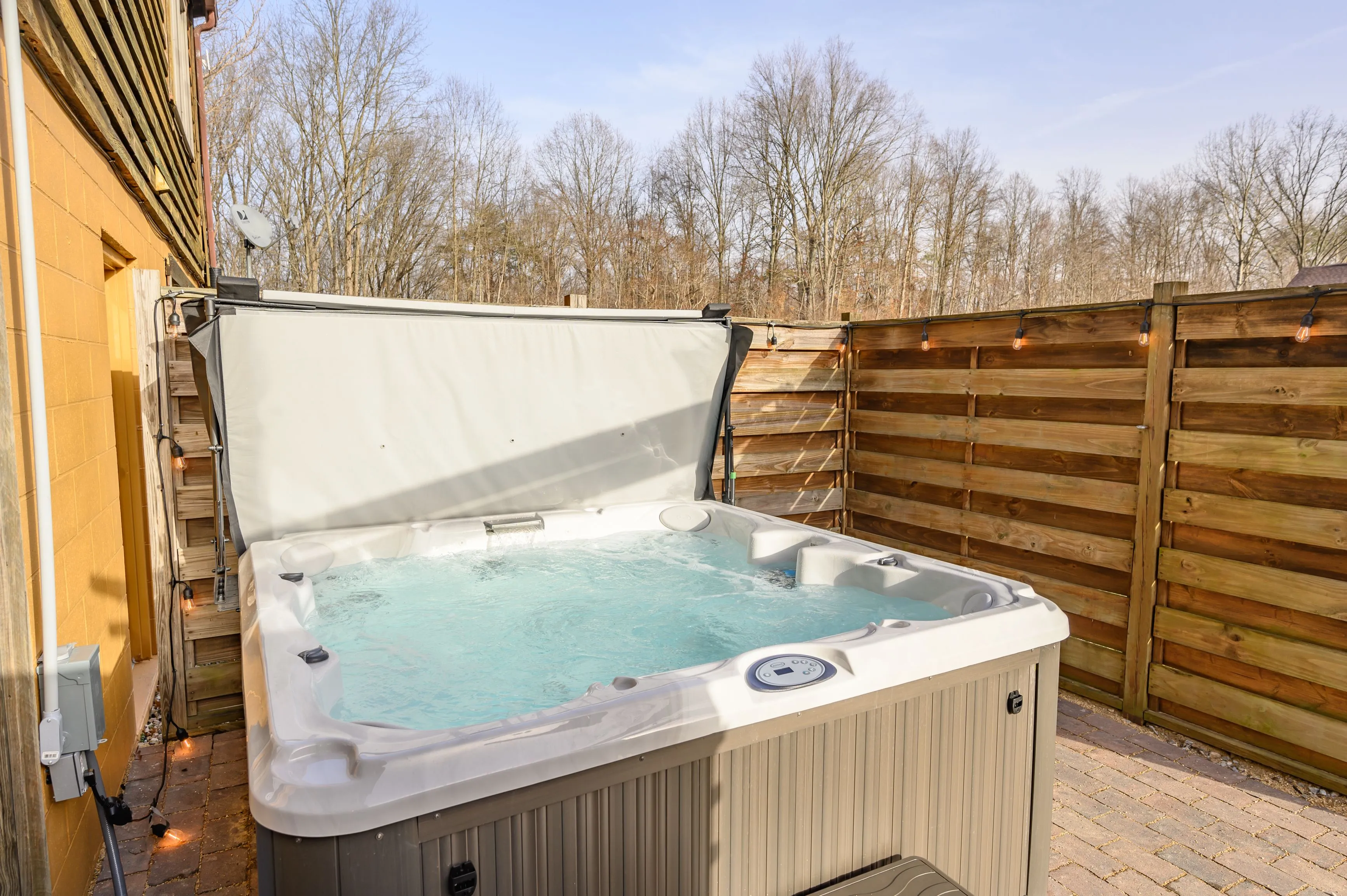 Outdoor home jacuzzi with open cover on a patio, surrounded by a wooden fence, with bare trees and blue sky in the background.