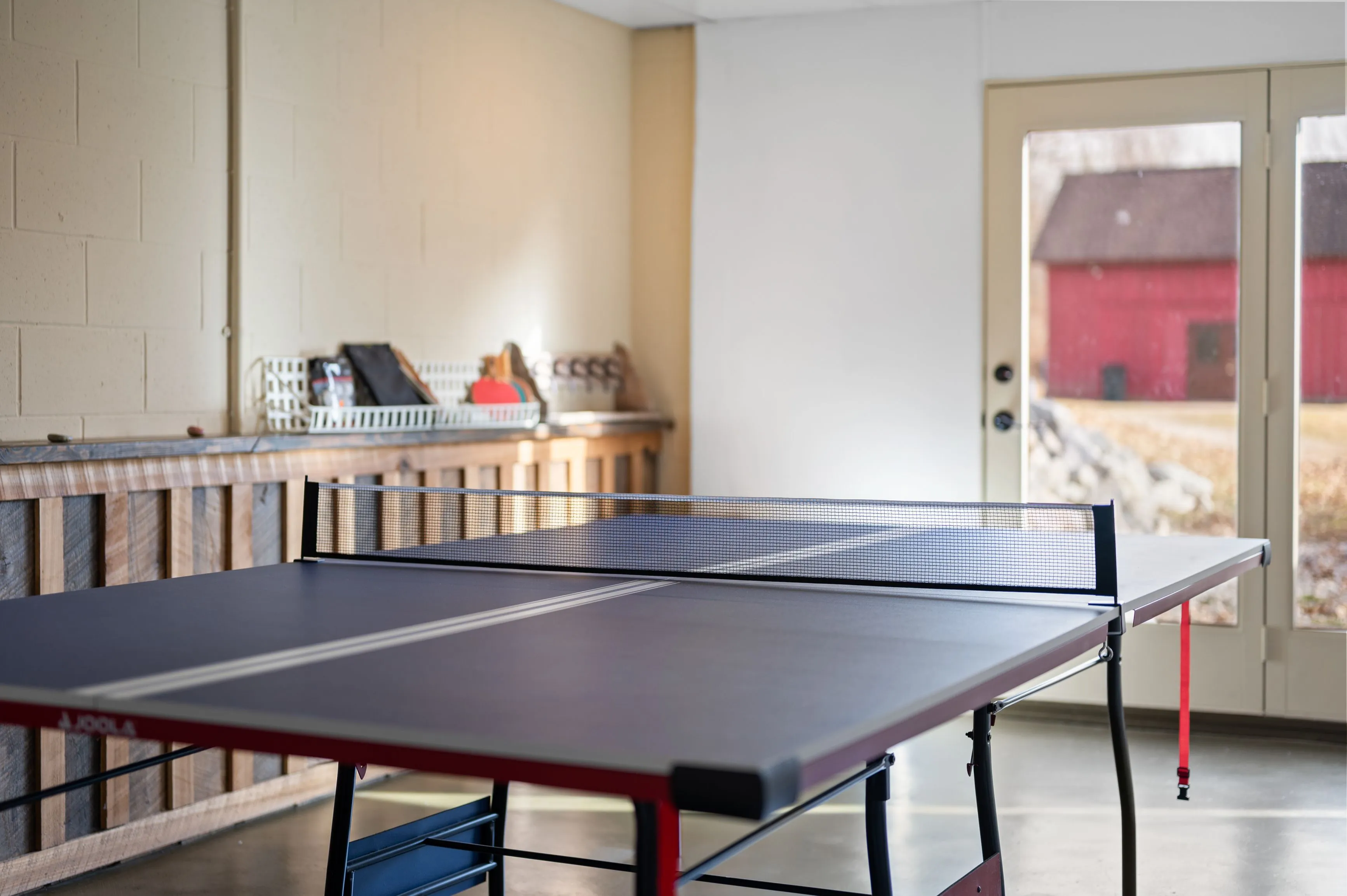 A table tennis table in a spacious room with concrete walls, a workbench with tools, and view of a red barn outside through the door.